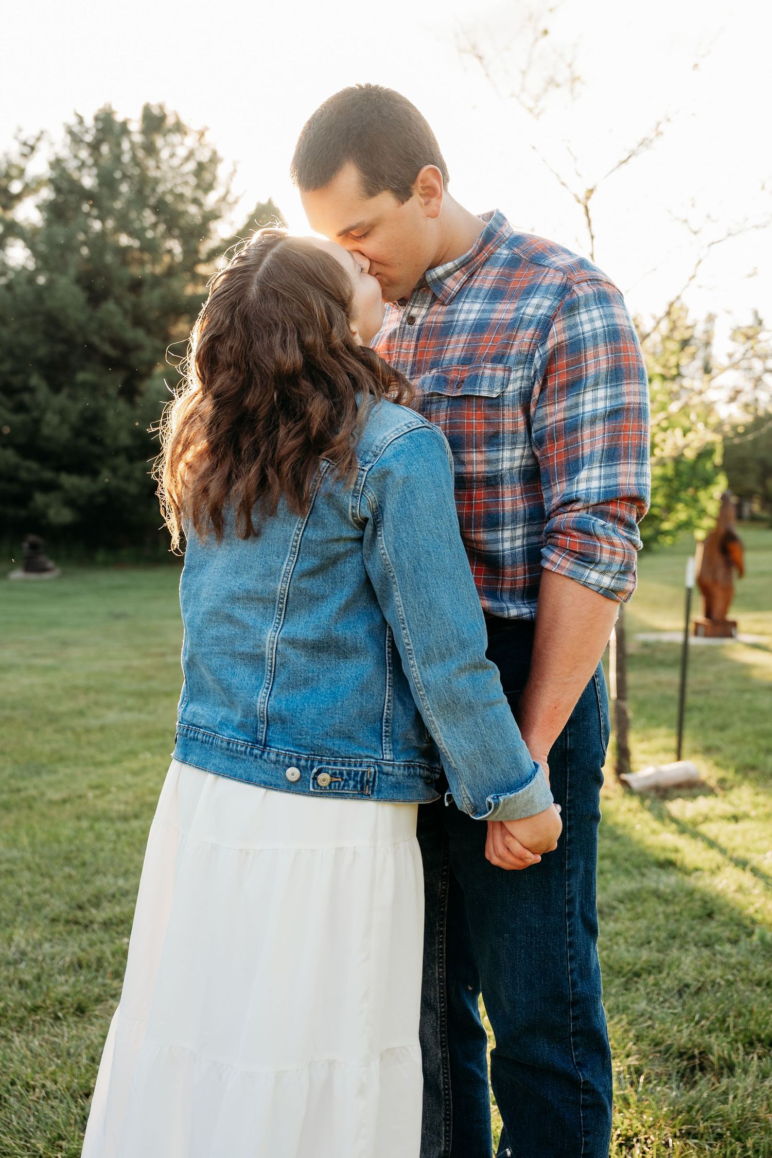 Couple sharing a tender kiss outdoors at golden hour, woman in denim jacket and white skirt holding hands with man in plaid shirt.
