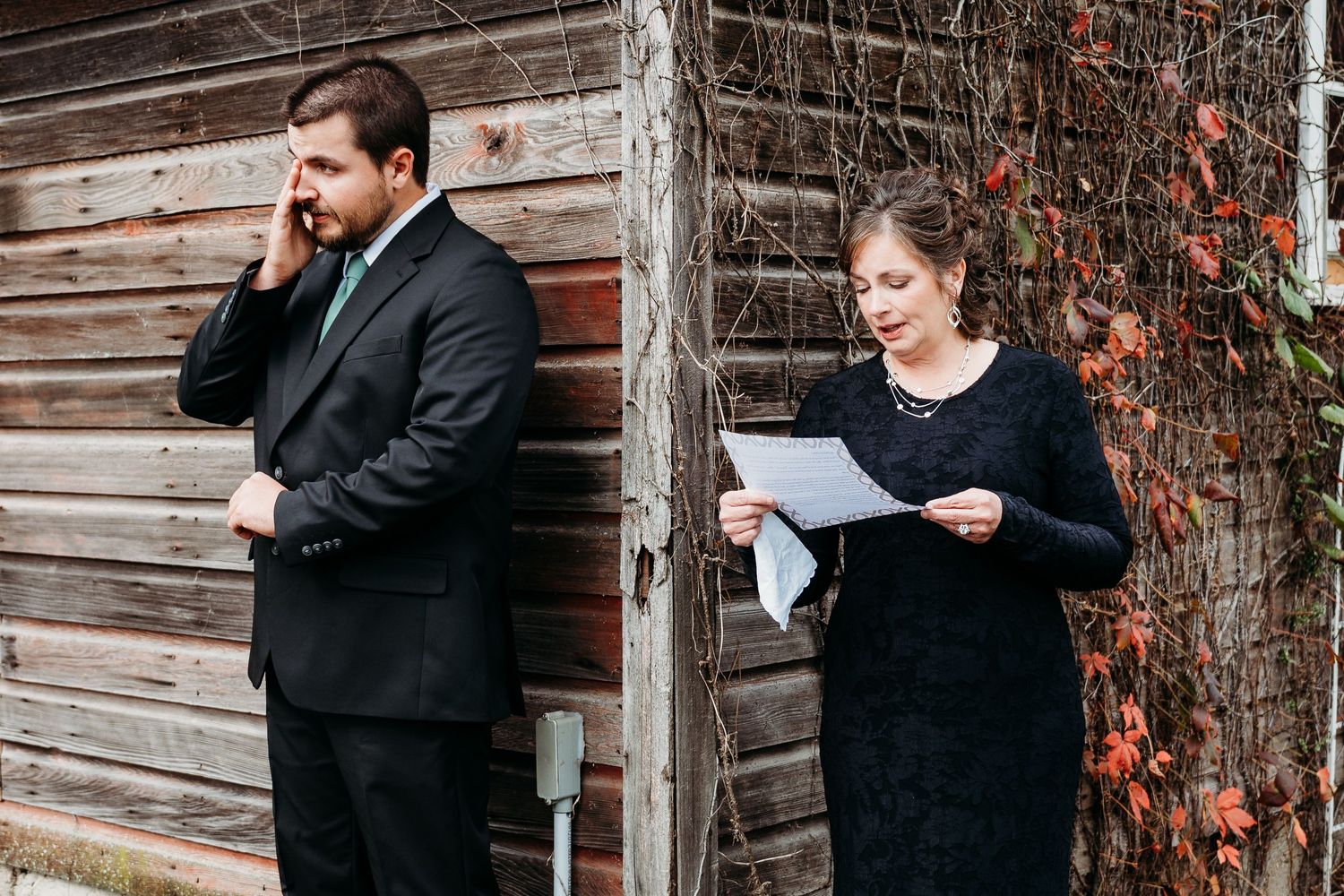 Two people in formal black attire stand against a rustic wooden wall with autumn ivy, one reading papers and one on phone.