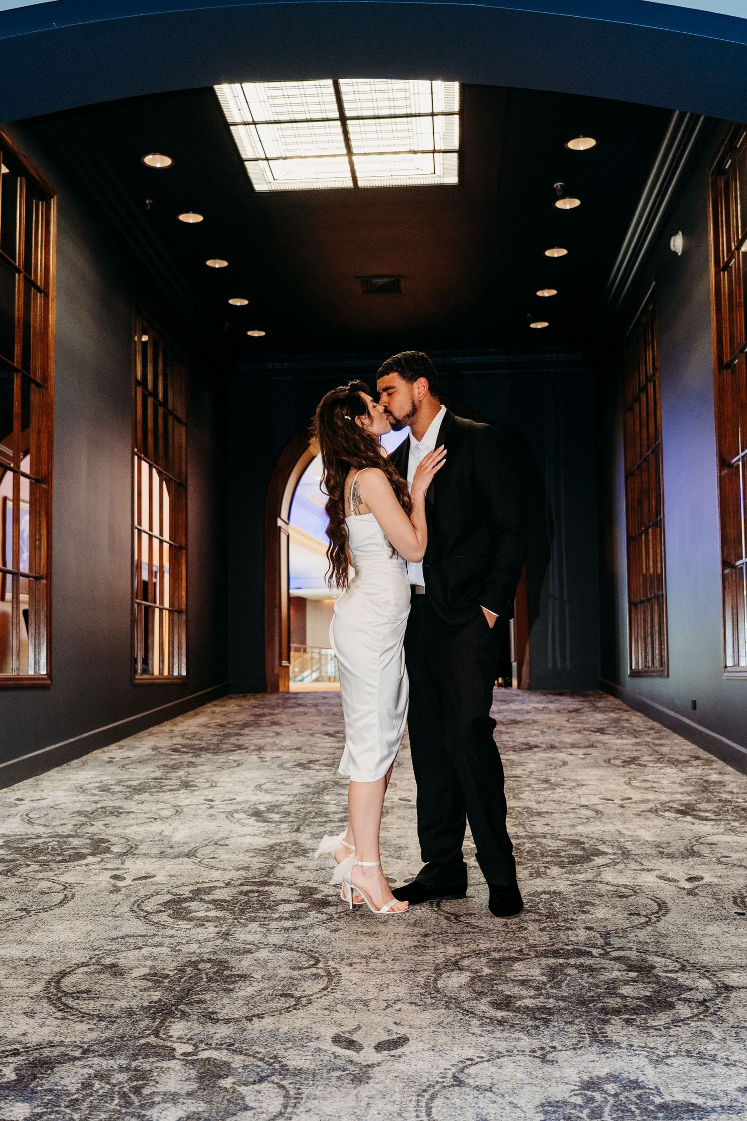 Couple embraces in romantic kiss within elegant dark hallway featuring ornate carpet and skylight lighting.