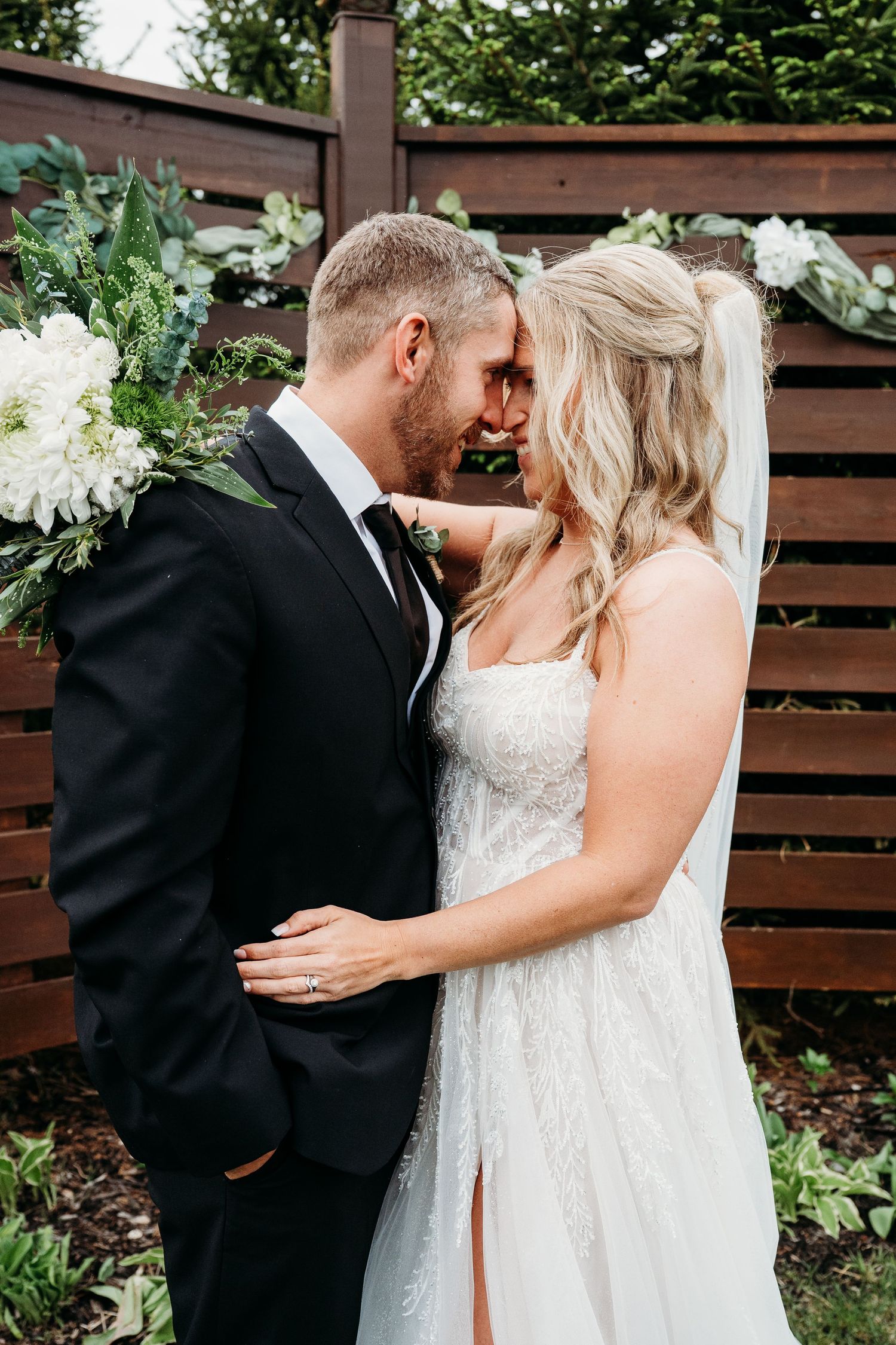 Wedding couple shares an intimate moment in front of a wooden fence with white floral decorations.