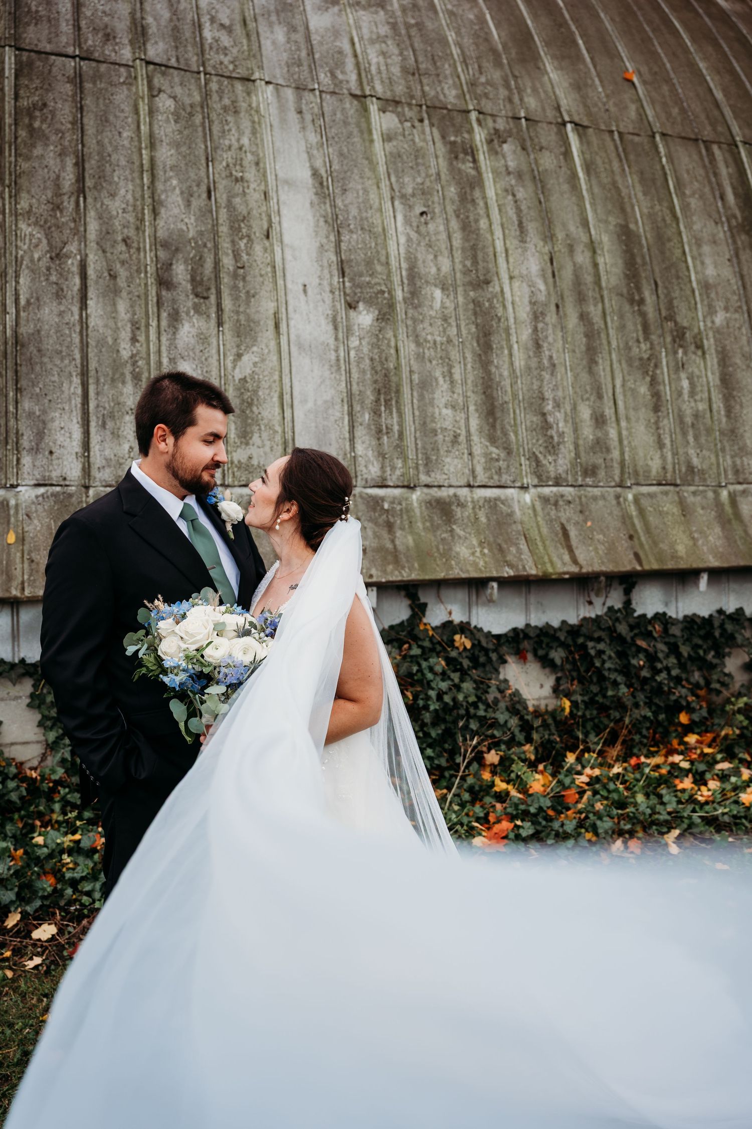 A romantic wedding couple poses against a rustic barn wall with orange flowers and flowing white veil in the background.