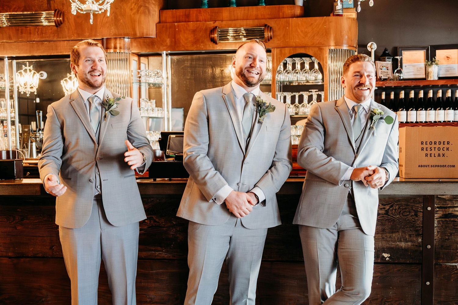 Wedding party members in light gray suits stand in sequence at a rustic bar venue.
