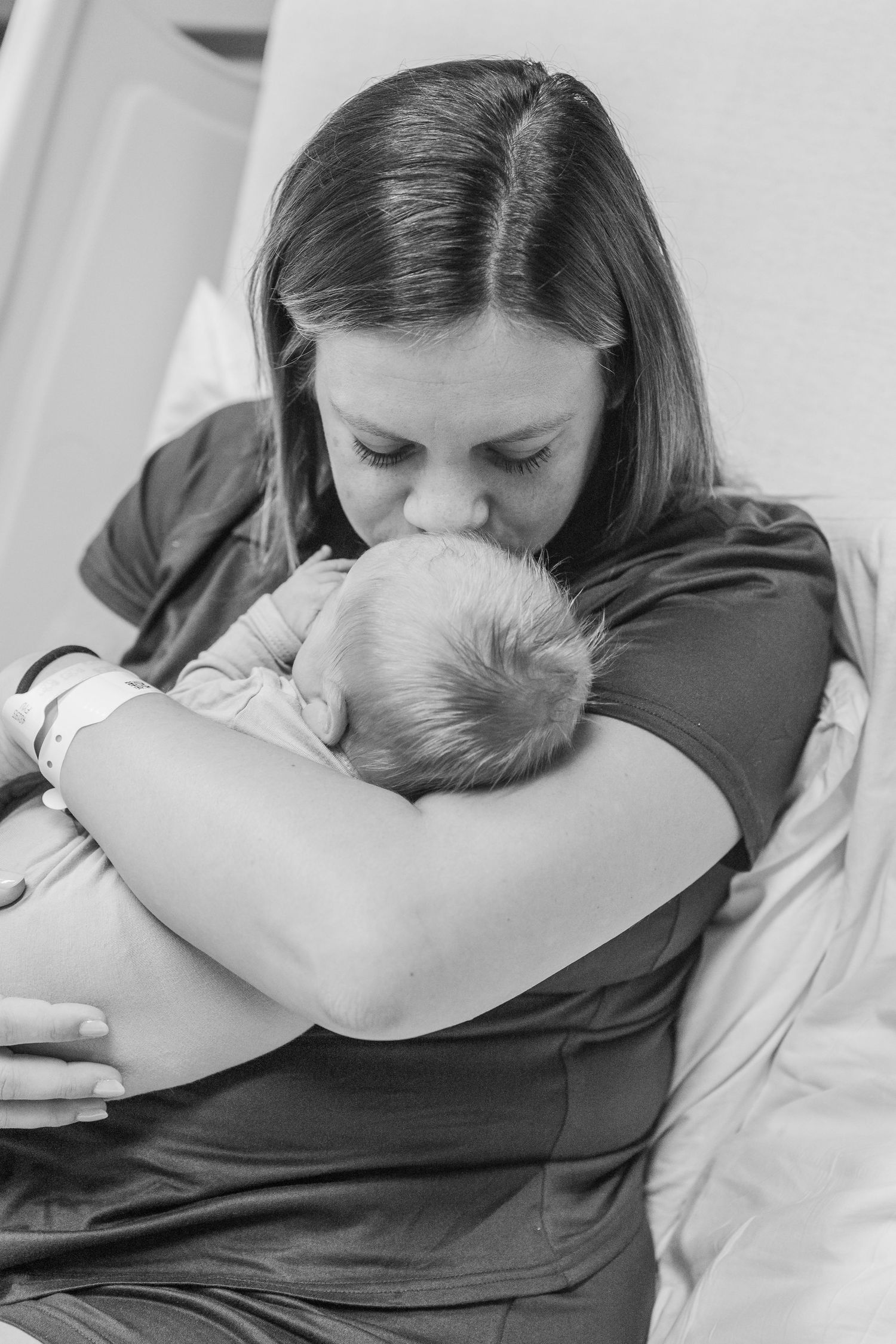 A tender black and white photograph showing a mother cradling and kissing her newborn baby in a hospital setting.