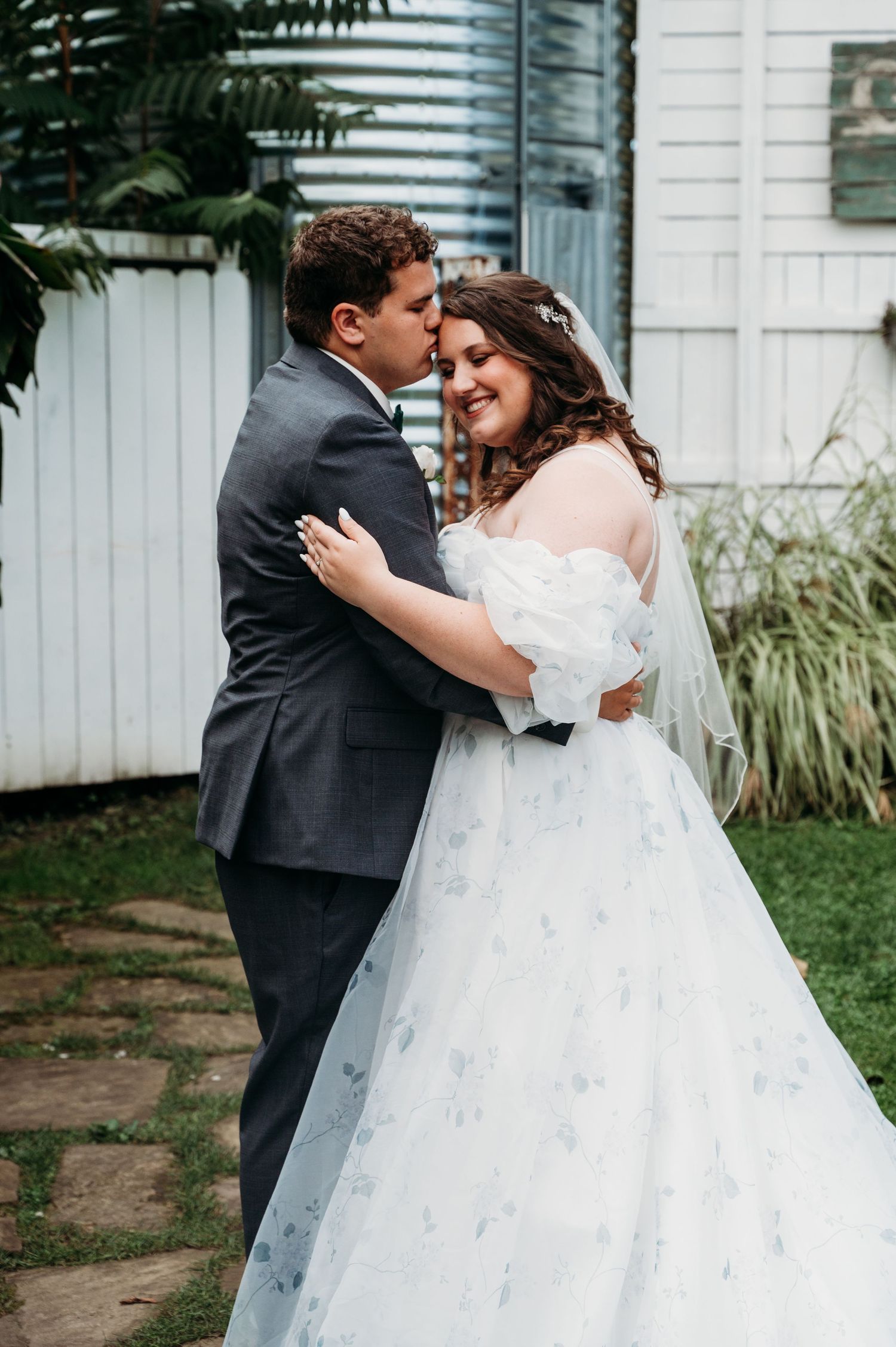 A couple shares a tender moment in their wedding attire in front of a white fence and greenery.