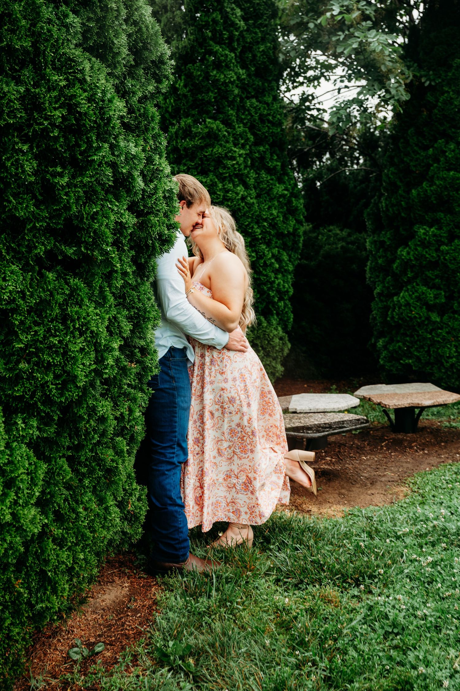 Couple sharing a tender kiss against tall green hedges in a serene garden setting with stone benches.