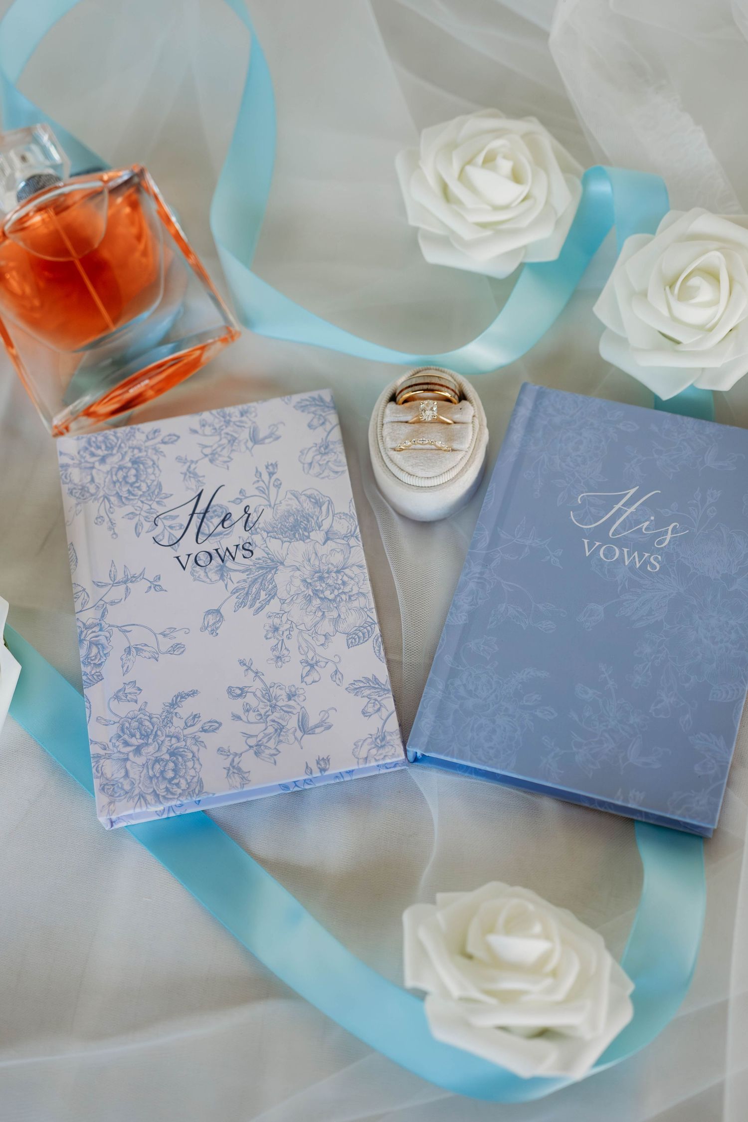 His and Her vows books with wedding rings, white roses, blue ribbon, and orange perfume bottle on white fabric.