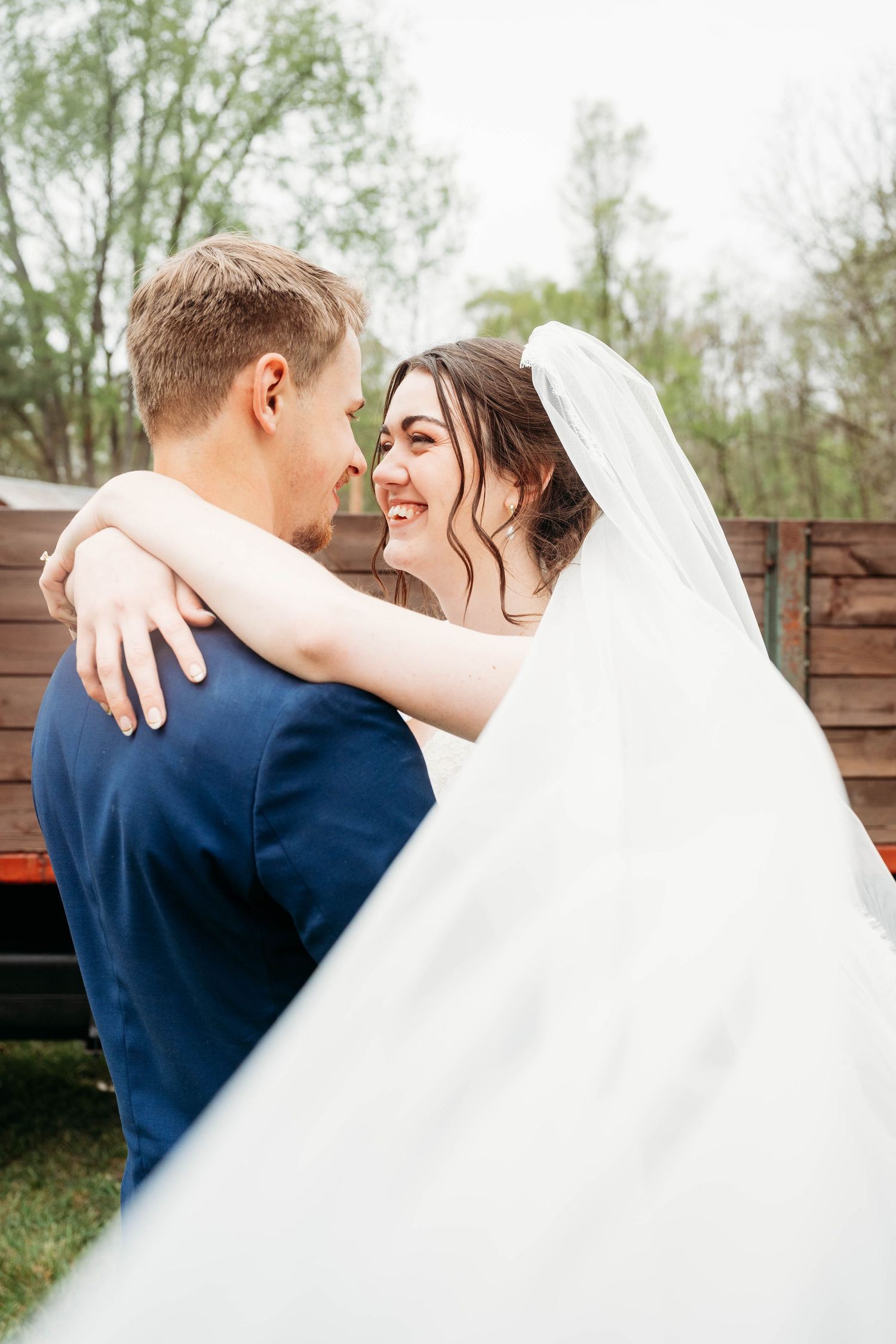 A joyful bride in a white veil embraces her groom in a navy suit outdoors at a rustic wedding venue.