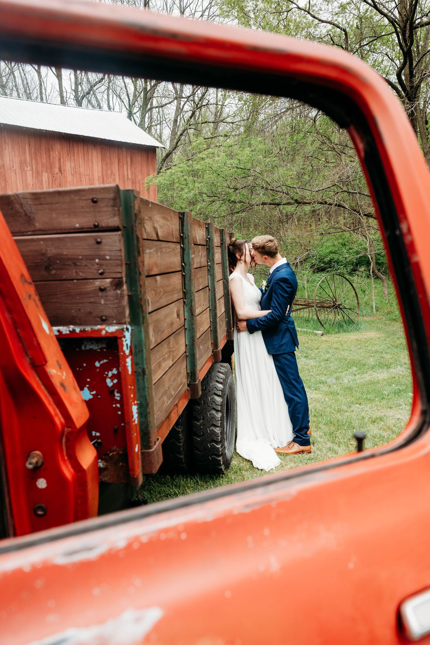 Bride and groom share a romantic moment beside a vintage red farm truck, viewed through the truck window.