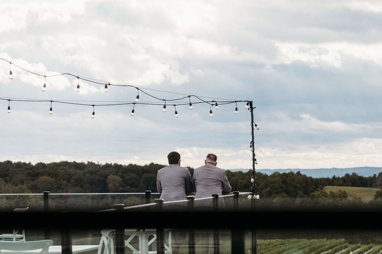 Two people sit together on a deck overlooking scenic countryside with string lights hanging overhead at dusk.