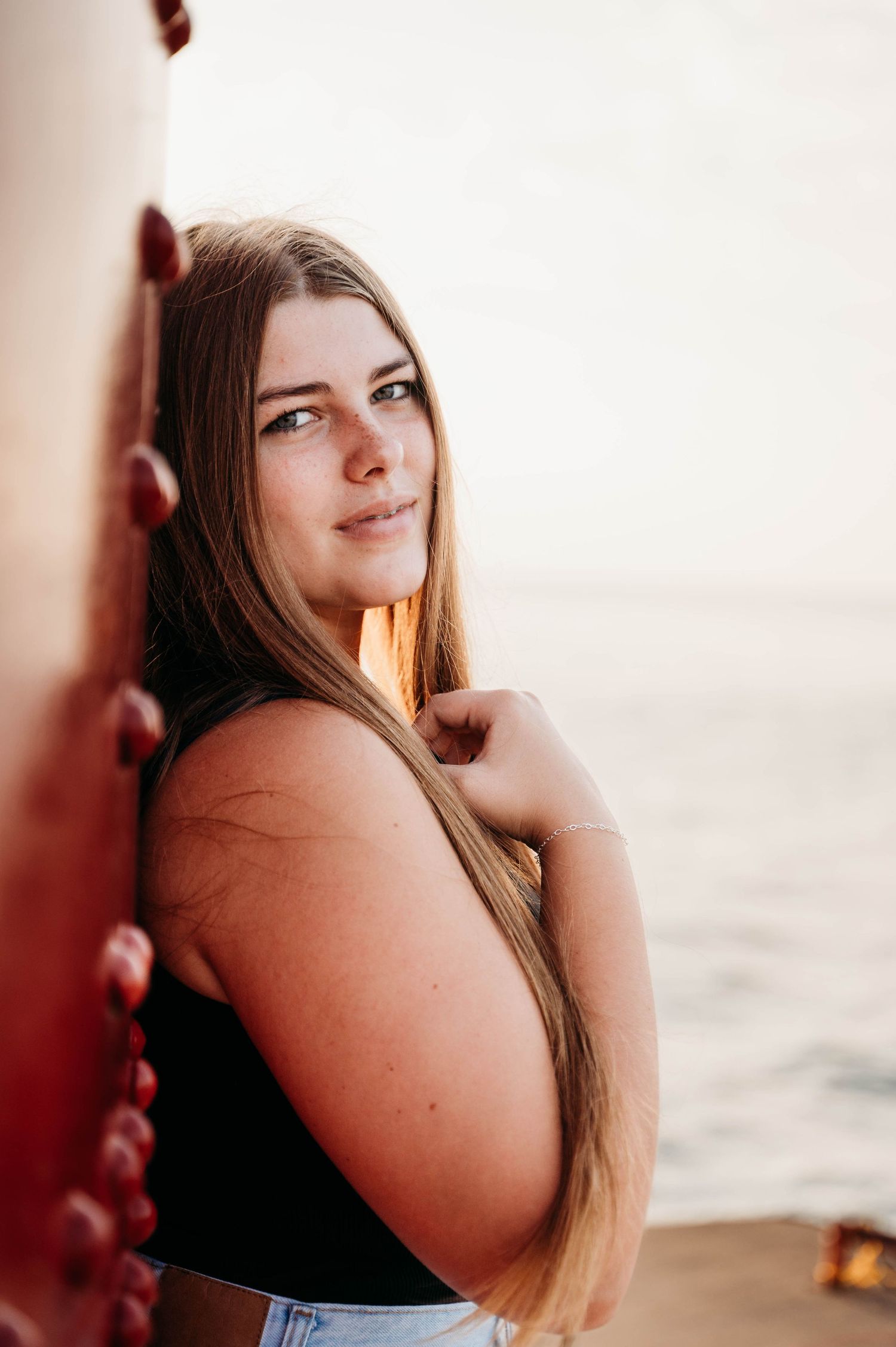 A young woman in a black top poses for a portrait by water during golden hour with warm natural lighting.
