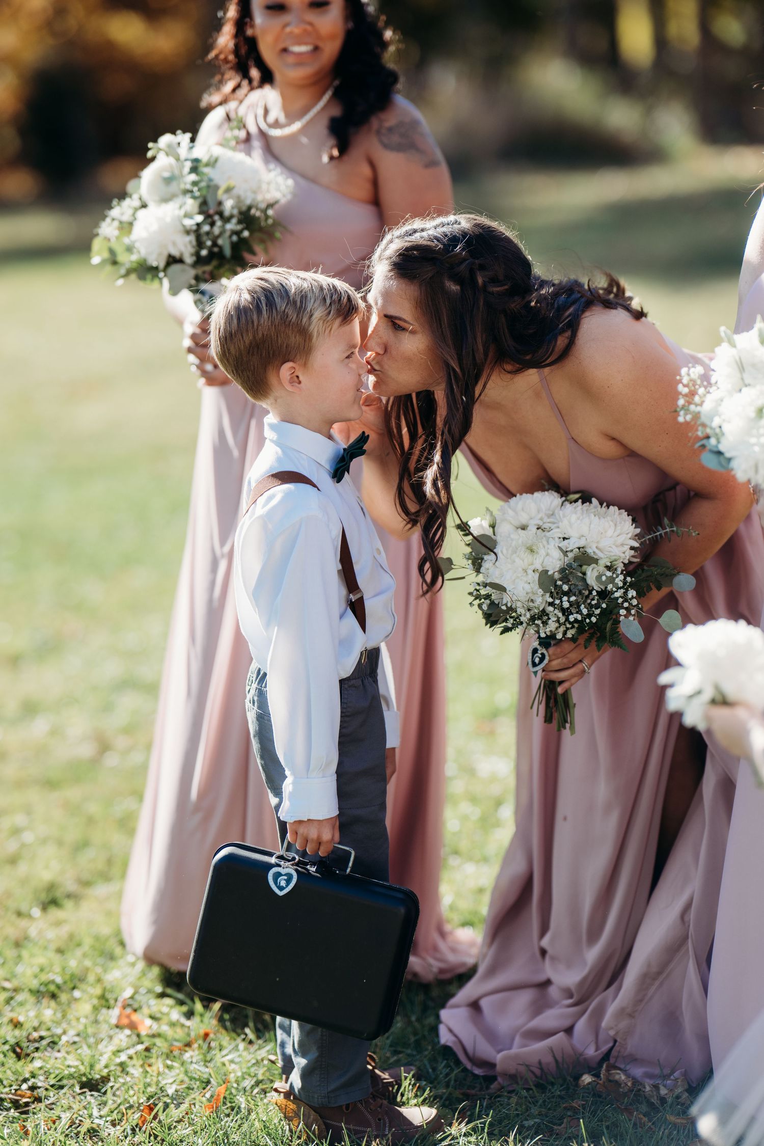Sweet moment at wedding as young ring bearer in suspenders shares tender moment with bridal party member in blush pink dress.