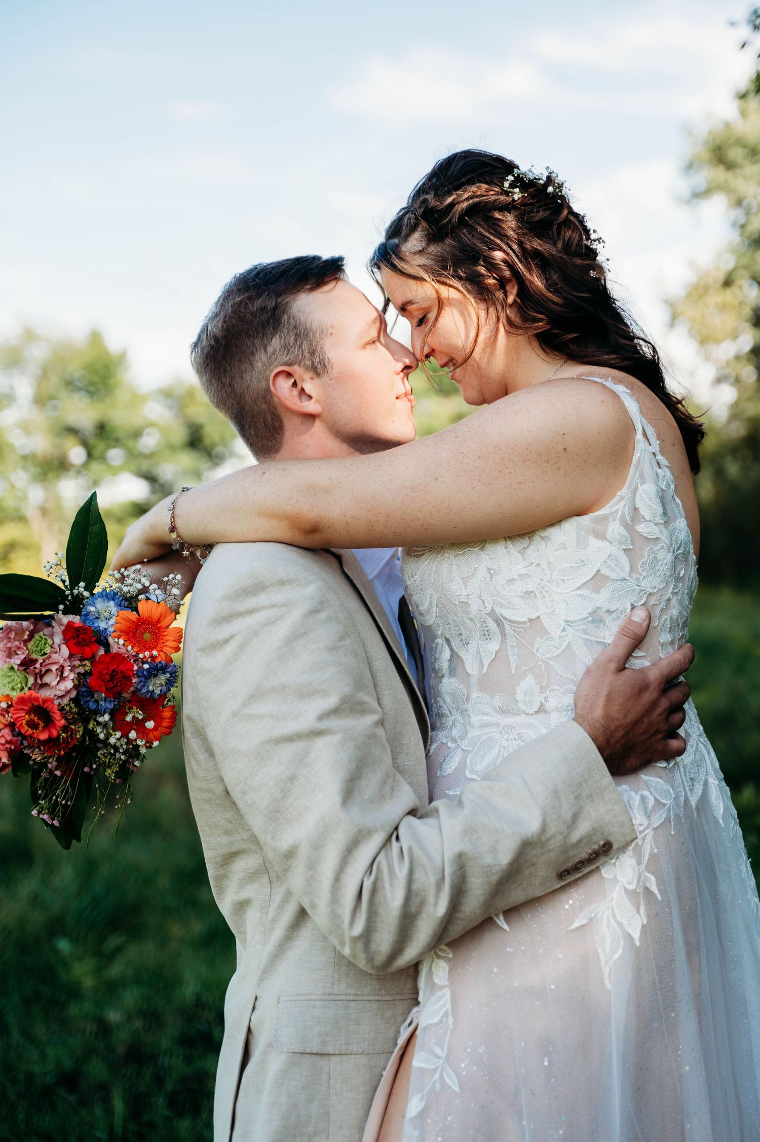 A romantic wedding moment outdoors with a couple embracing while the bride holds a vibrant red and orange bouquet.