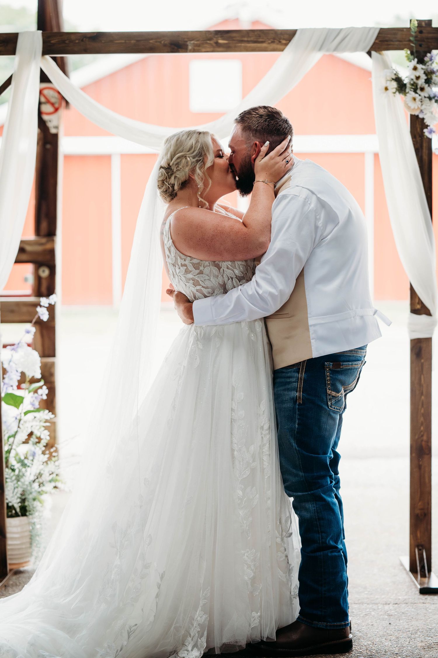 A couple shares a romantic kiss during their wedding ceremony under a wooden arch with flowing white drapery and pink backdrop.