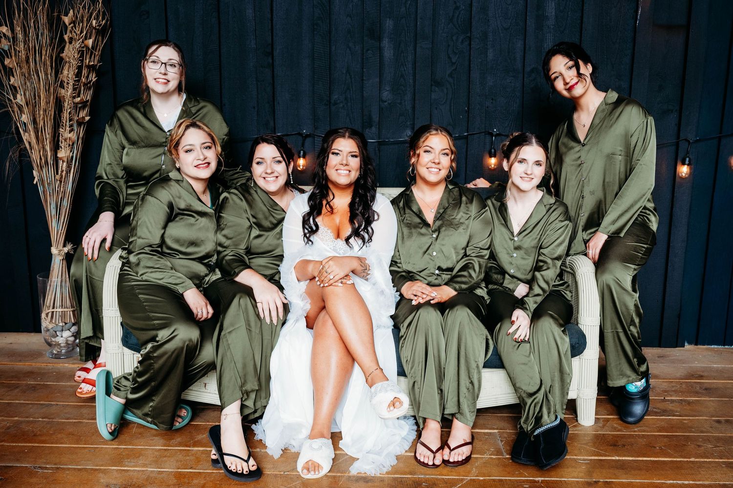 Bride in white gown poses with six bridesmaids wearing matching olive green satin robes on a white couch.
