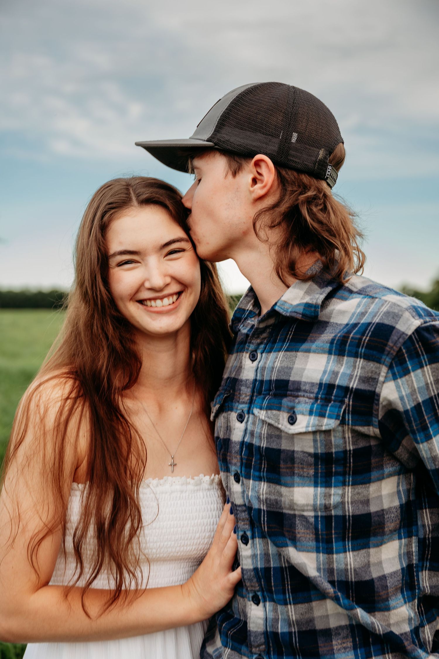 A young couple sharing a tender moment outdoors, man in plaid shirt kissing smiling woman in white dress.