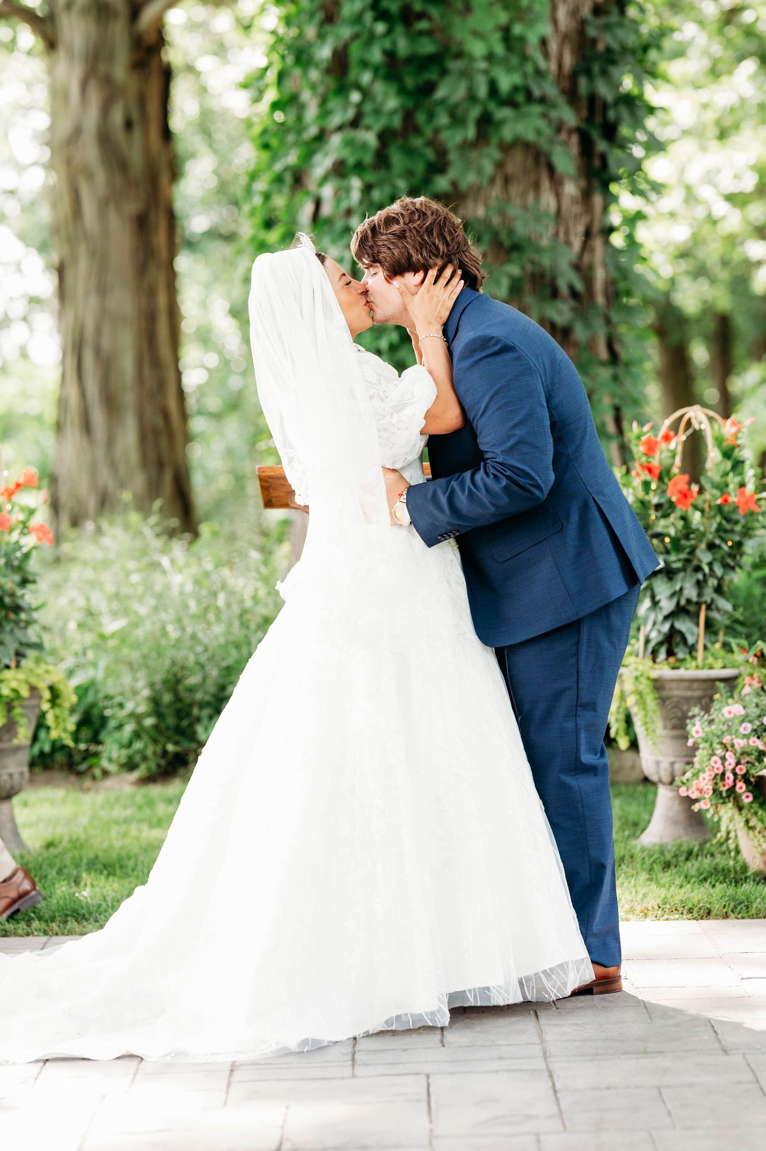 Bride and groom share first kiss outdoors, bride in white ball gown with veil, groom in navy suit.