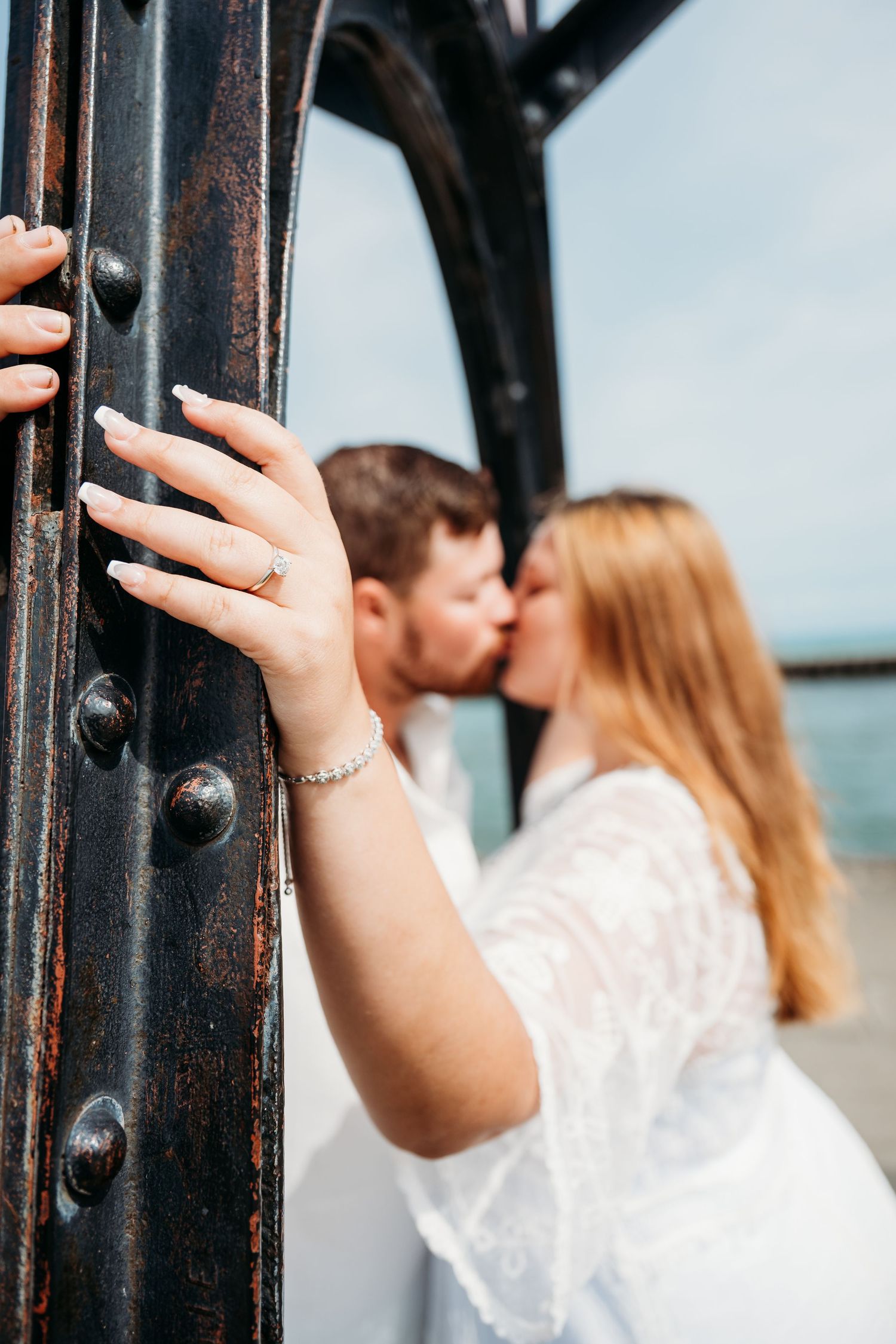 A romantic couple sharing a kiss with a woman's hand displaying an engagement ring against a metal structure.