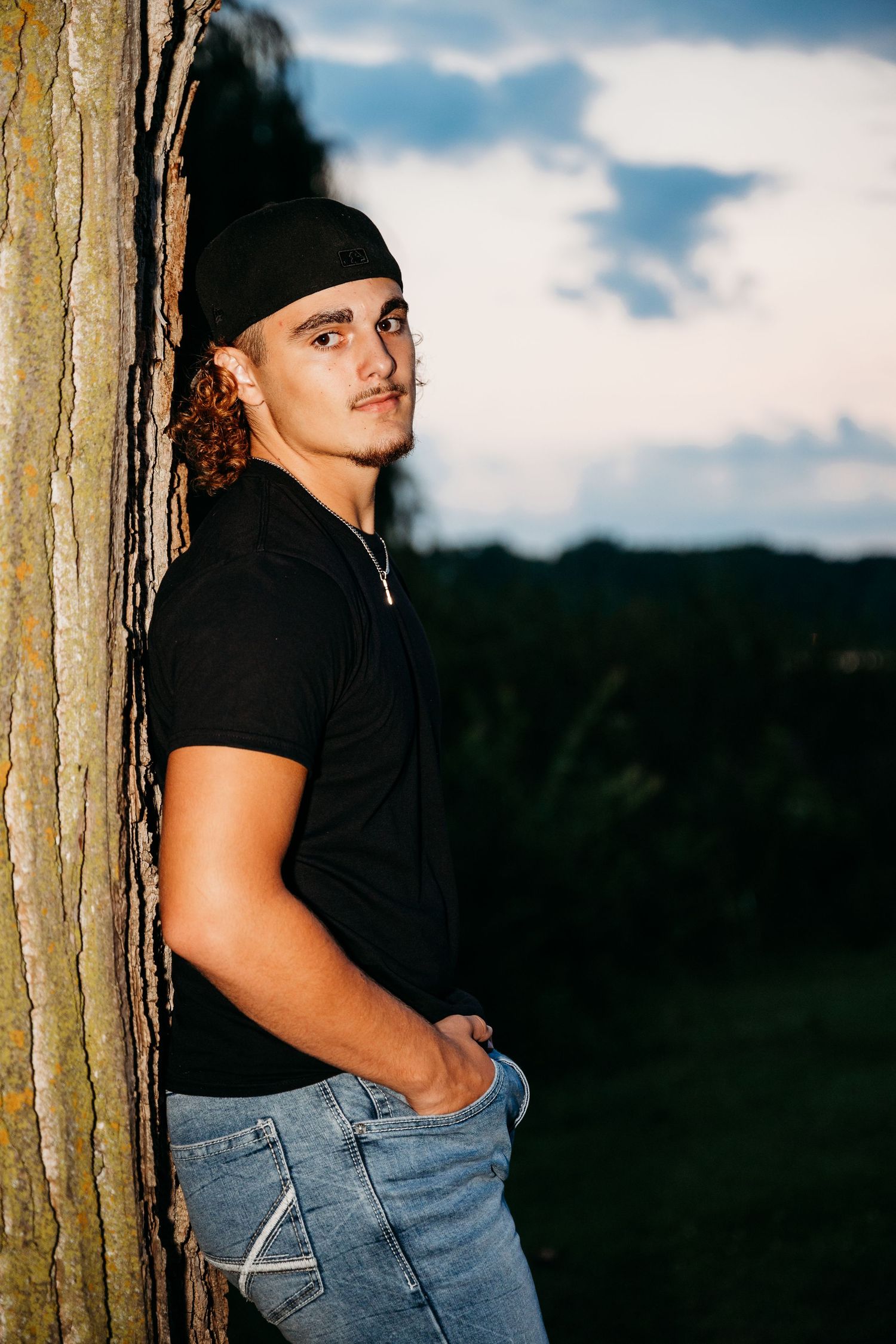 Person in black top and jeans poses against tree trunk at sunset with muted natural landscape in background.