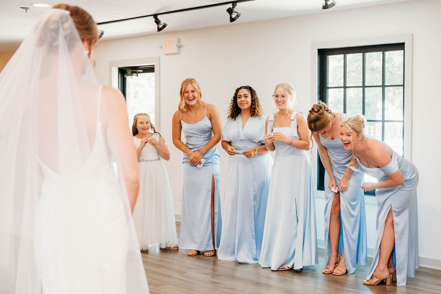 Bridesmaids in light blue dresses react with joy and excitement as bride reveals her wedding gown during bridal party getting ready.