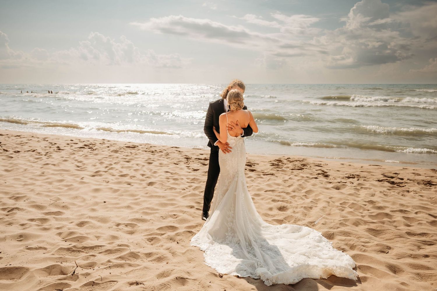 Bride and groom embrace on sandy beach with ocean waves, bride wearing elegant lace wedding gown with long train.