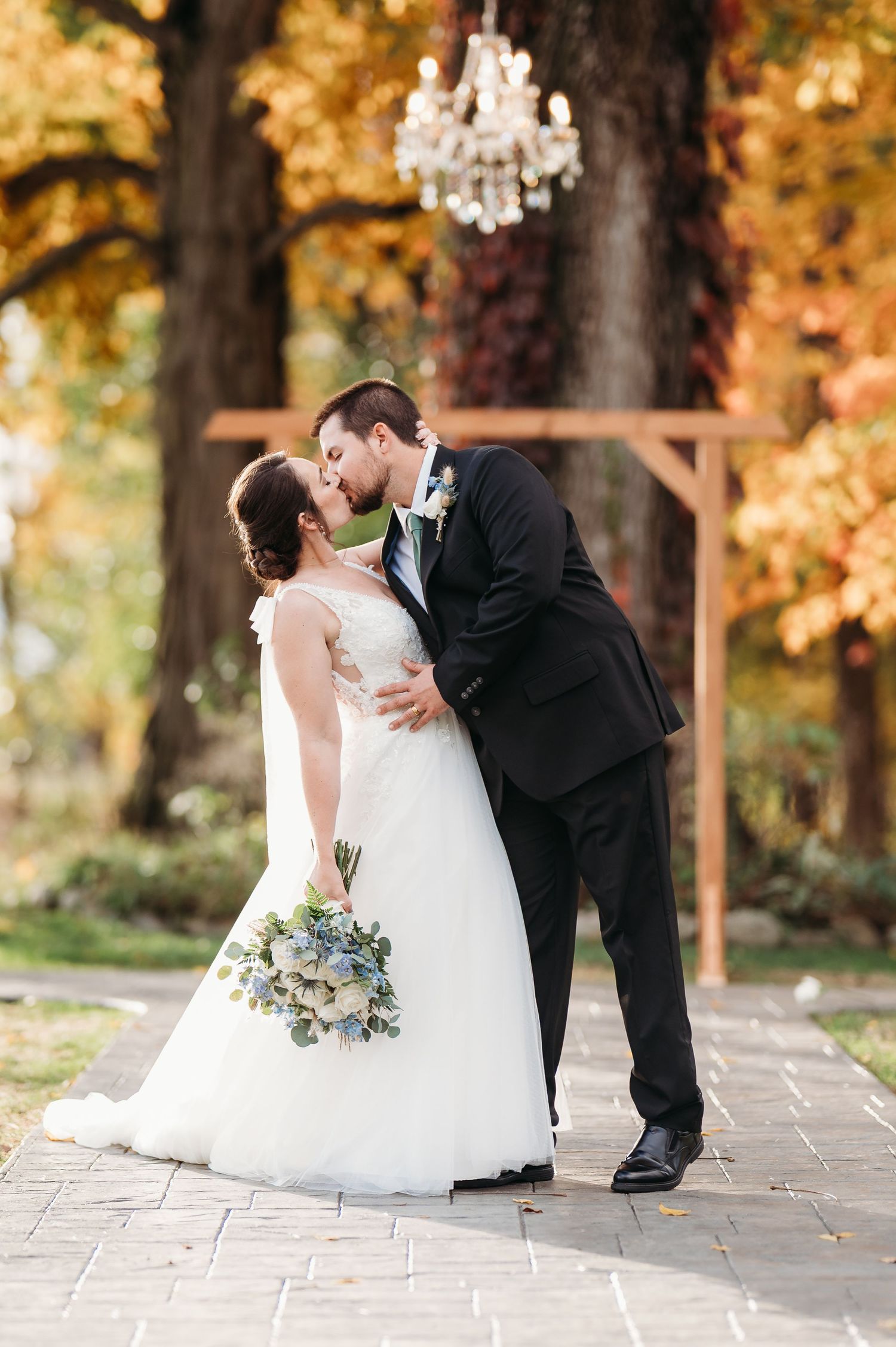 Bride and groom share a kiss outdoors in autumn, she holds a blue bouquet, wooden arch and chandelier behind them. Vintage Rose Barn
