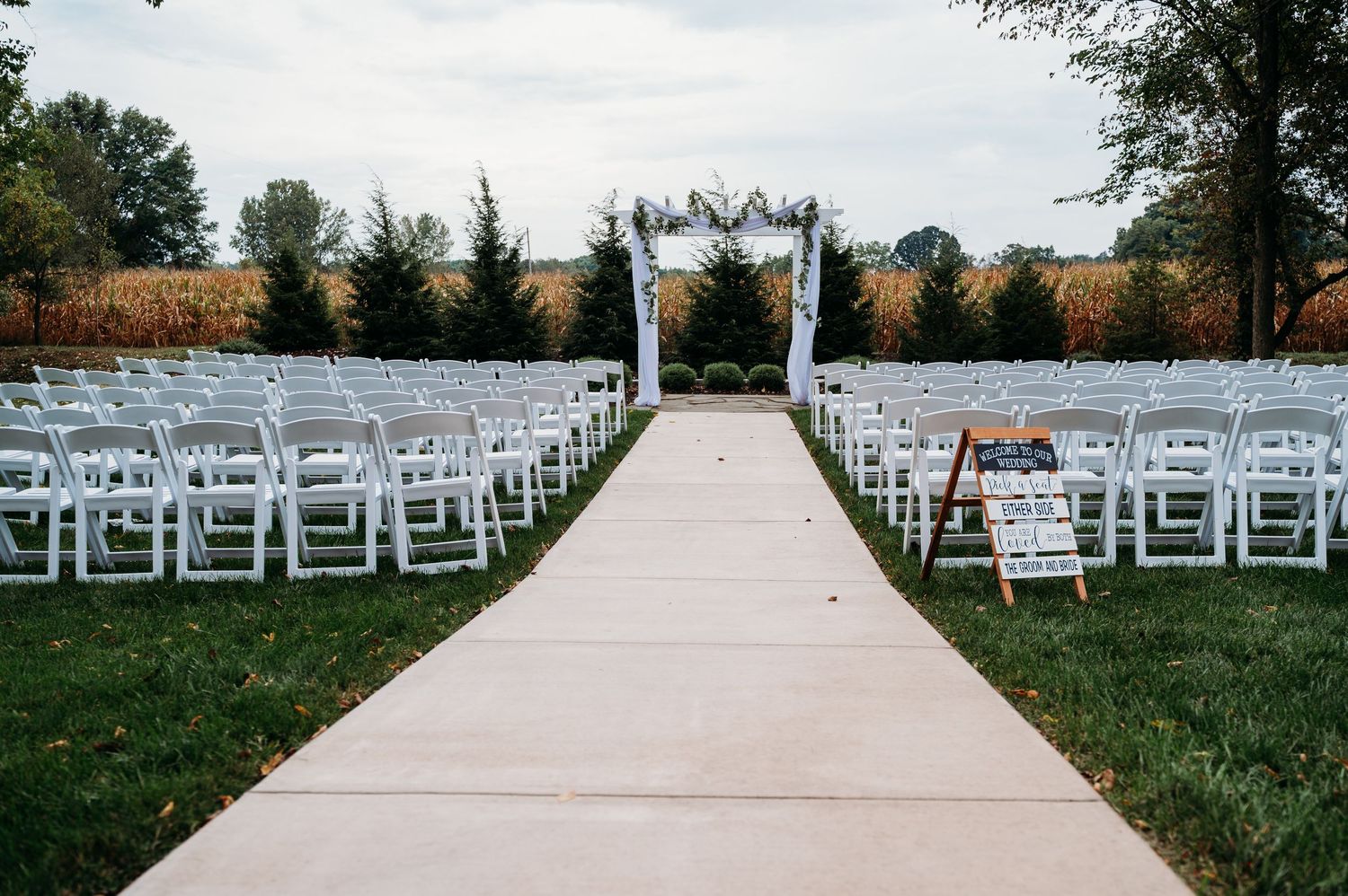Outdoor wedding ceremony setup with white folding chairs lining an aisle leading to a floral arch, surrounded by lush greenery.