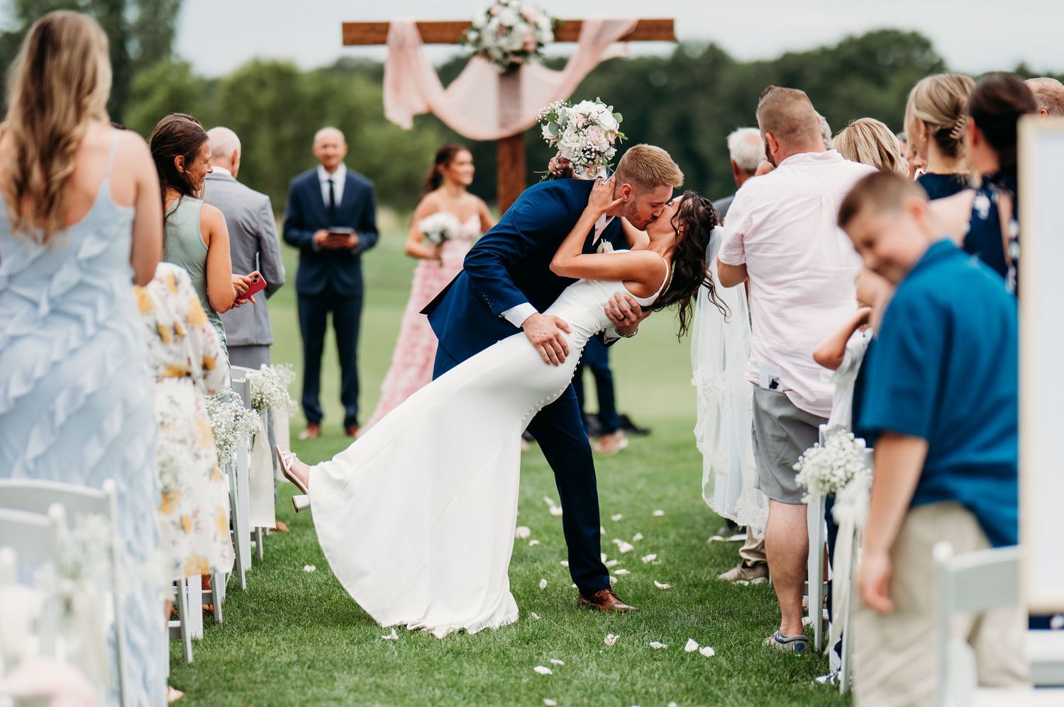 Groom dips bride for a romantic kiss at outdoor wedding ceremony aisle surrounded by guests on green lawn.