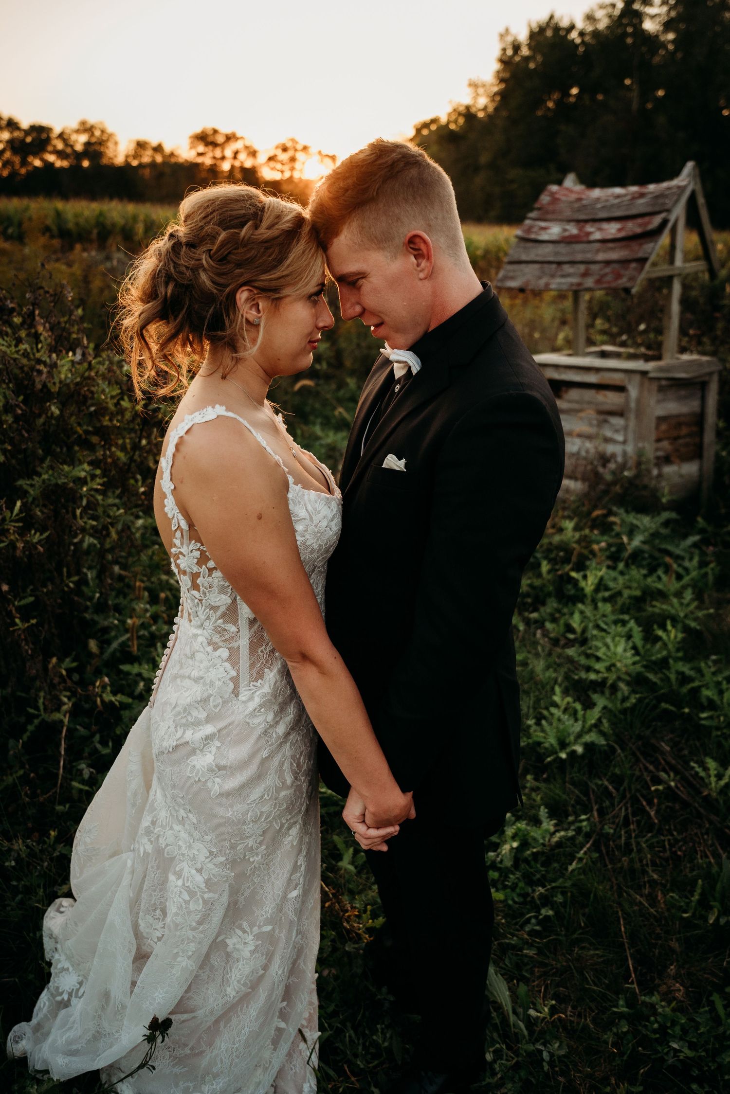 Bride and groom hold hands forehead-to-forehead at golden hour sunset in lush green outdoor wedding portrait.