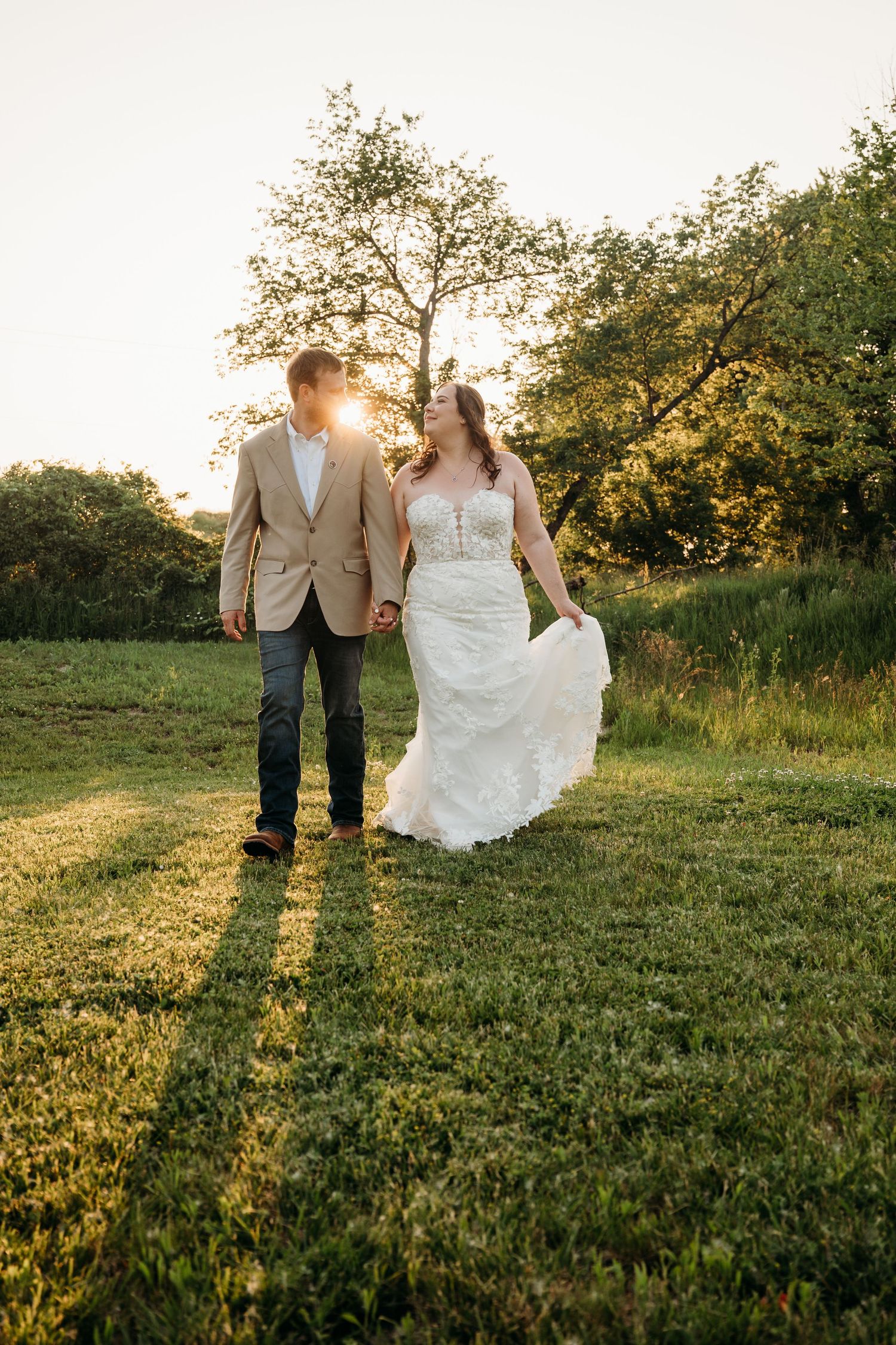 A bride and groom walk hand-in-hand through a green field at golden hour, with the sun glowing behind them.