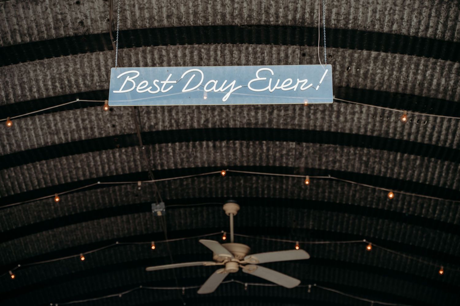 A decorative sign reading Best Day Ever hangs from a rustic barn ceiling with string lights and a ceiling fan.