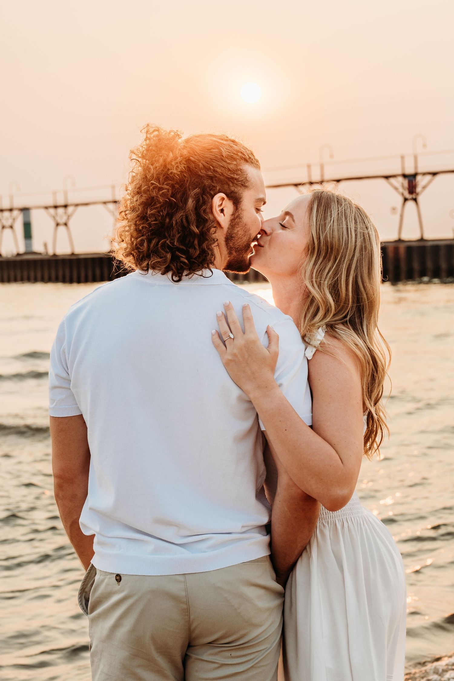 Couple sharing a romantic kiss at sunset by the water, with a pier in the background and warm golden light.