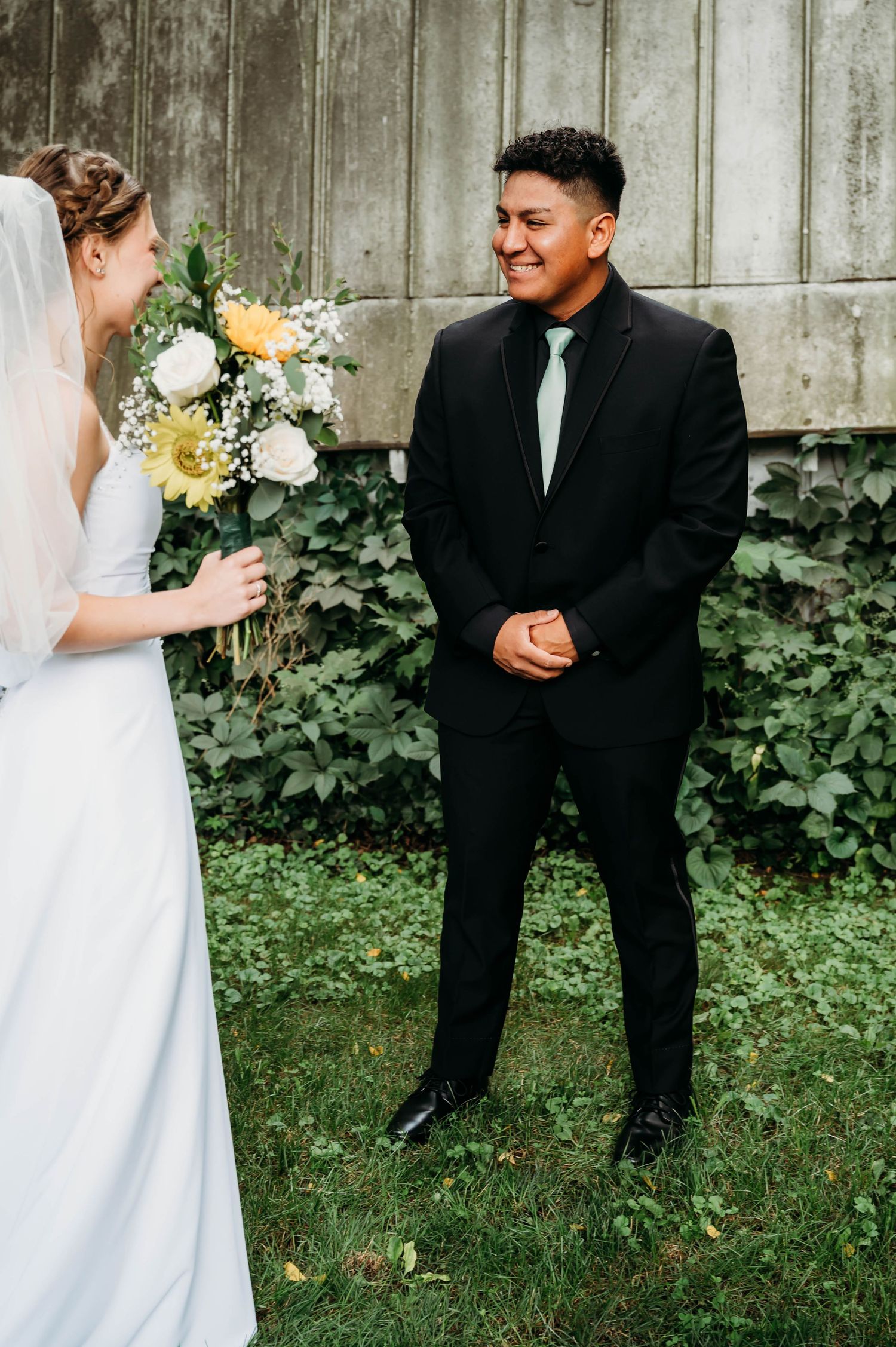 Groom in black suit smiling at bride holding yellow and white bouquet during outdoor first look wedding moment.