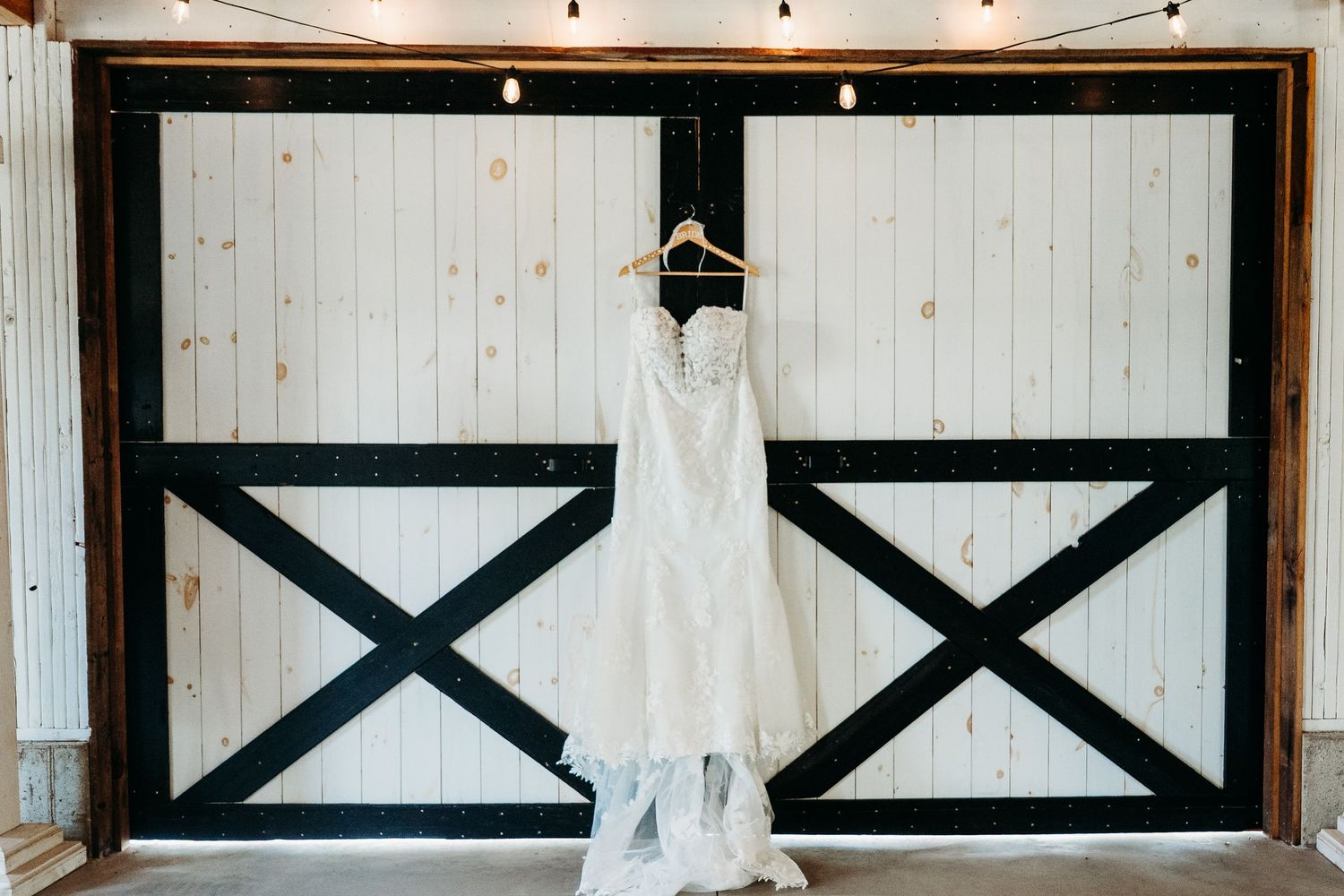 White wedding dress hanging on rustic barn door with black X pattern details and industrial lighting above.