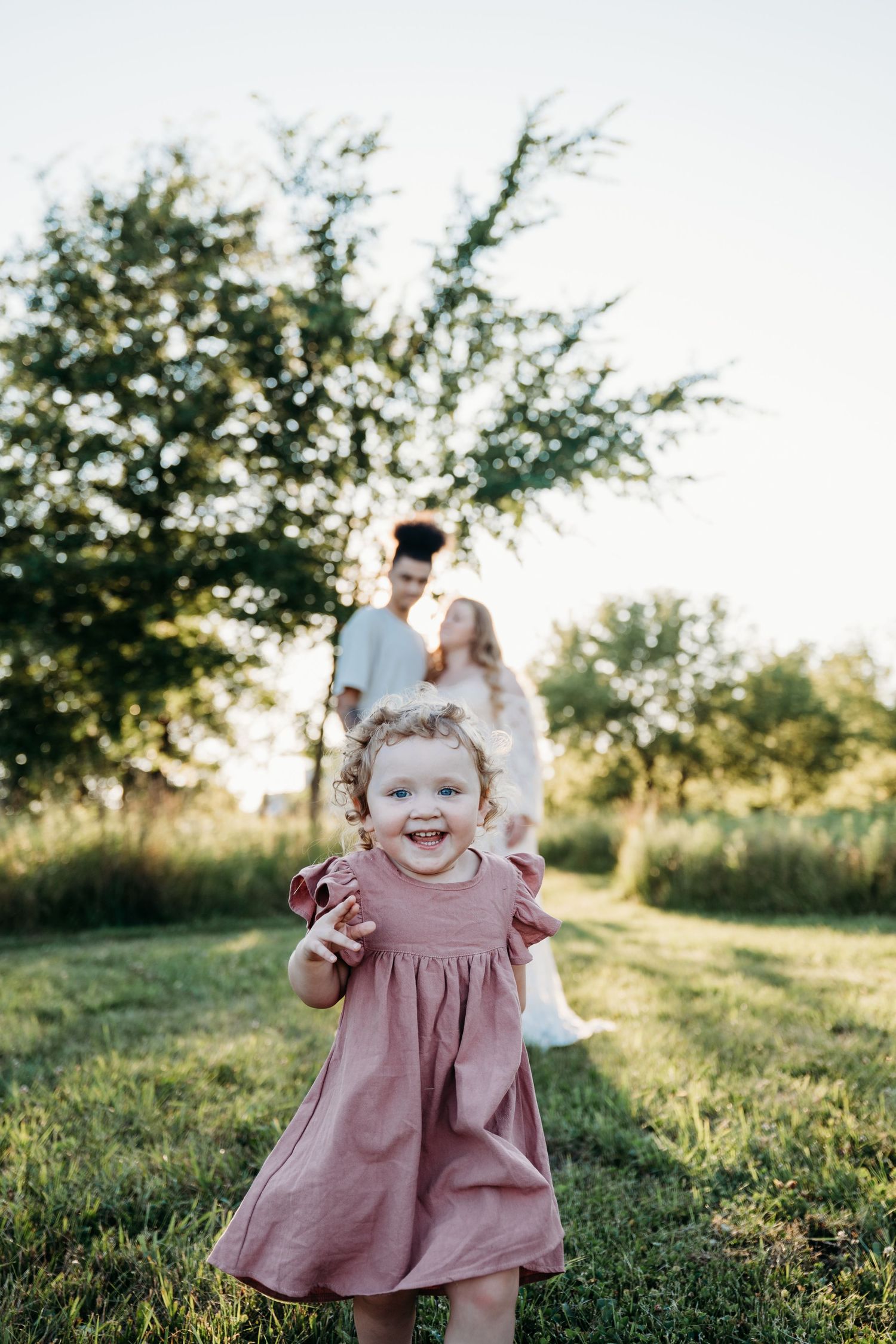 A blissful family portrait capturing tender moments between parents and a smiling toddler in a pink dress at golden hour.