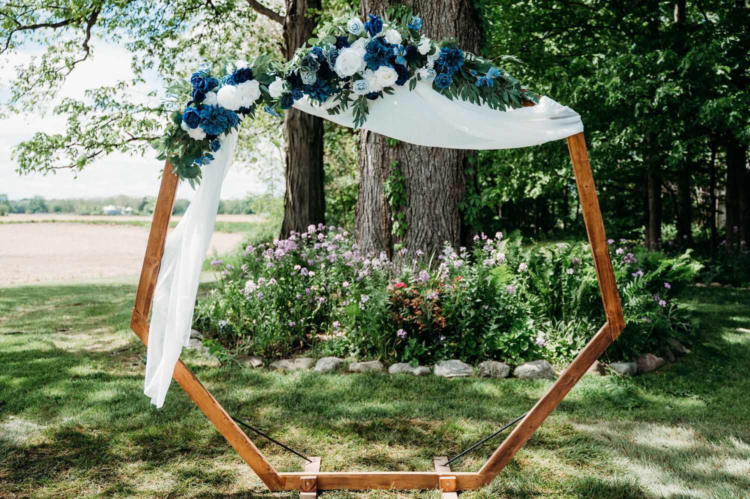 Modern hexagonal wooden wedding arch decorated with white fabric and blue flowers in an outdoor garden setting.