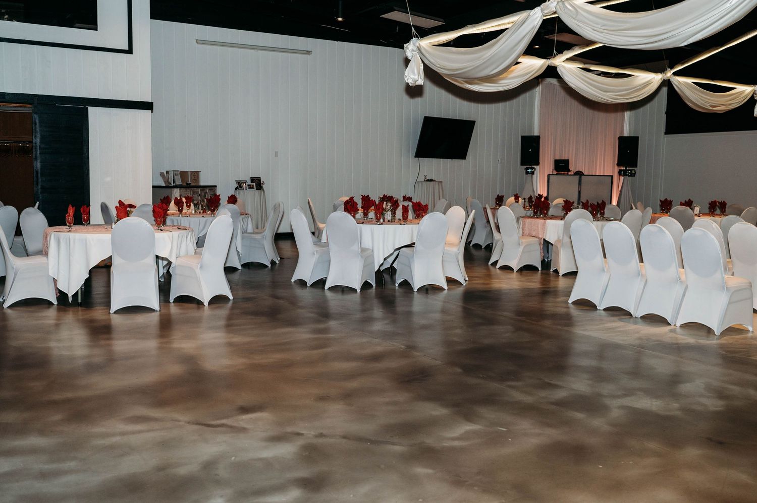 White fabric chair covers and elegant draping decorate an empty wedding reception venue with hardwood floors.