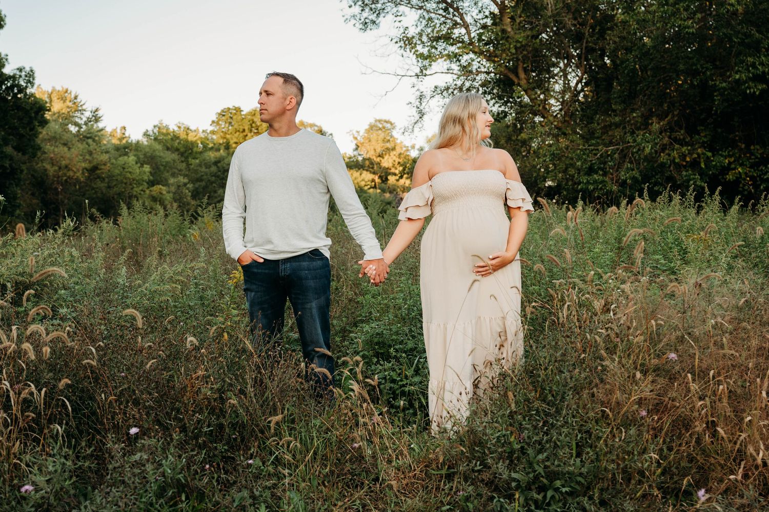 A couple walks through a tall grass meadow during golden hour while holding hands and wearing light colored clothing.