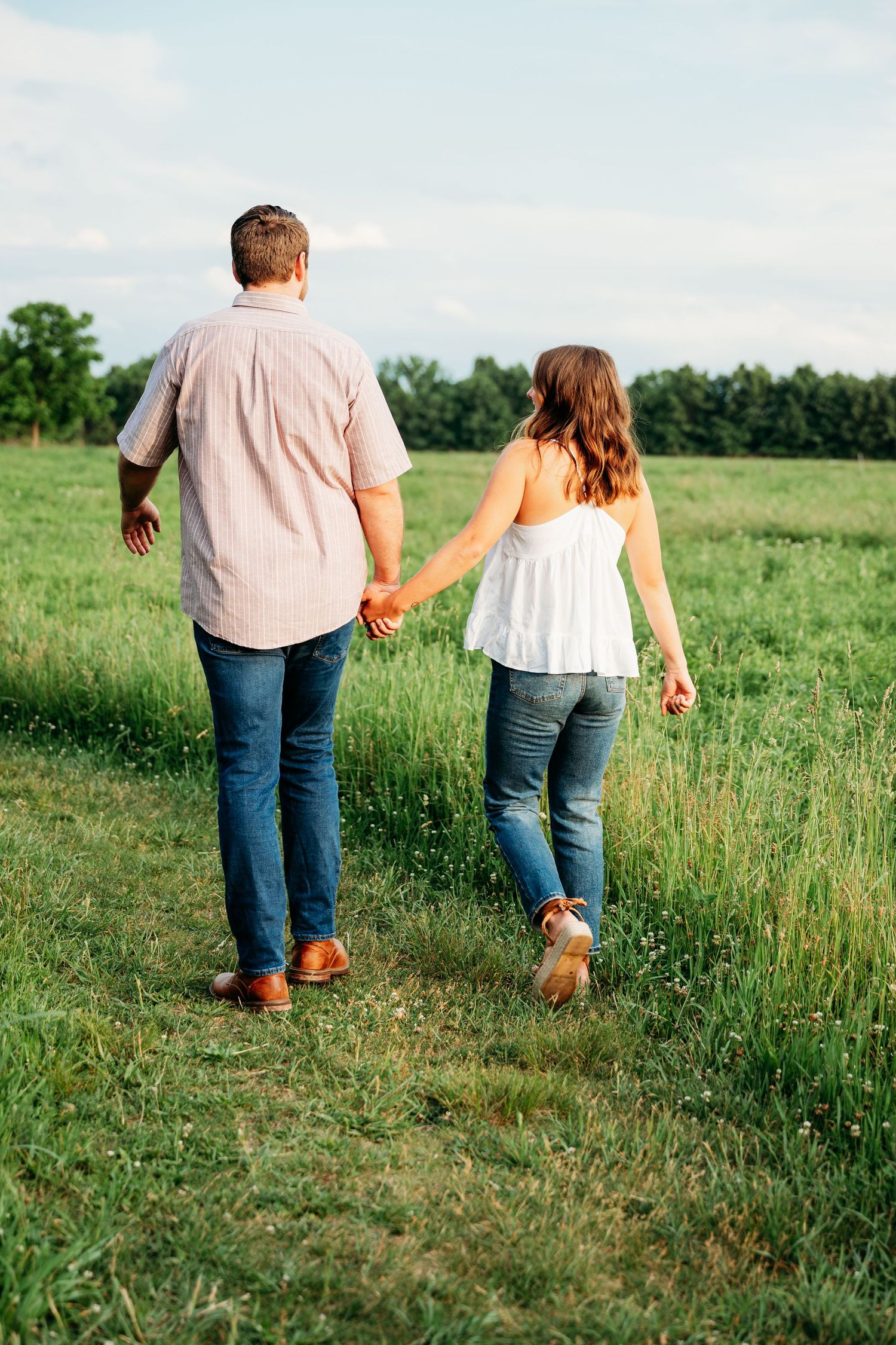 Couple holding hands walking through a lush green field, man in pink shirt and woman in white top, both wearing jeans.