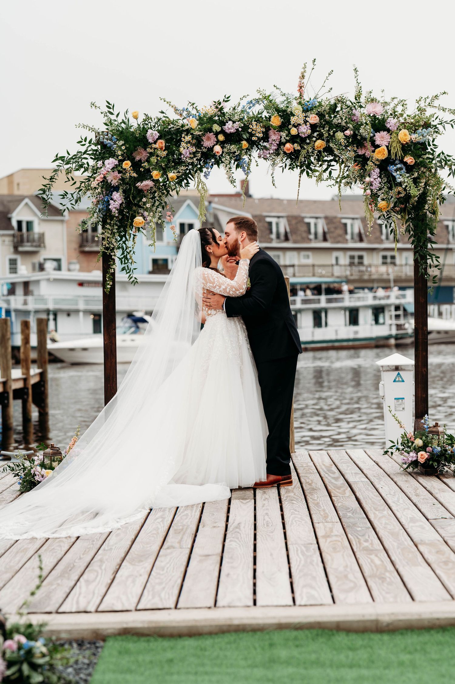 Bride and groom share first kiss under floral arch on waterfront dock at outdoor wedding ceremony.