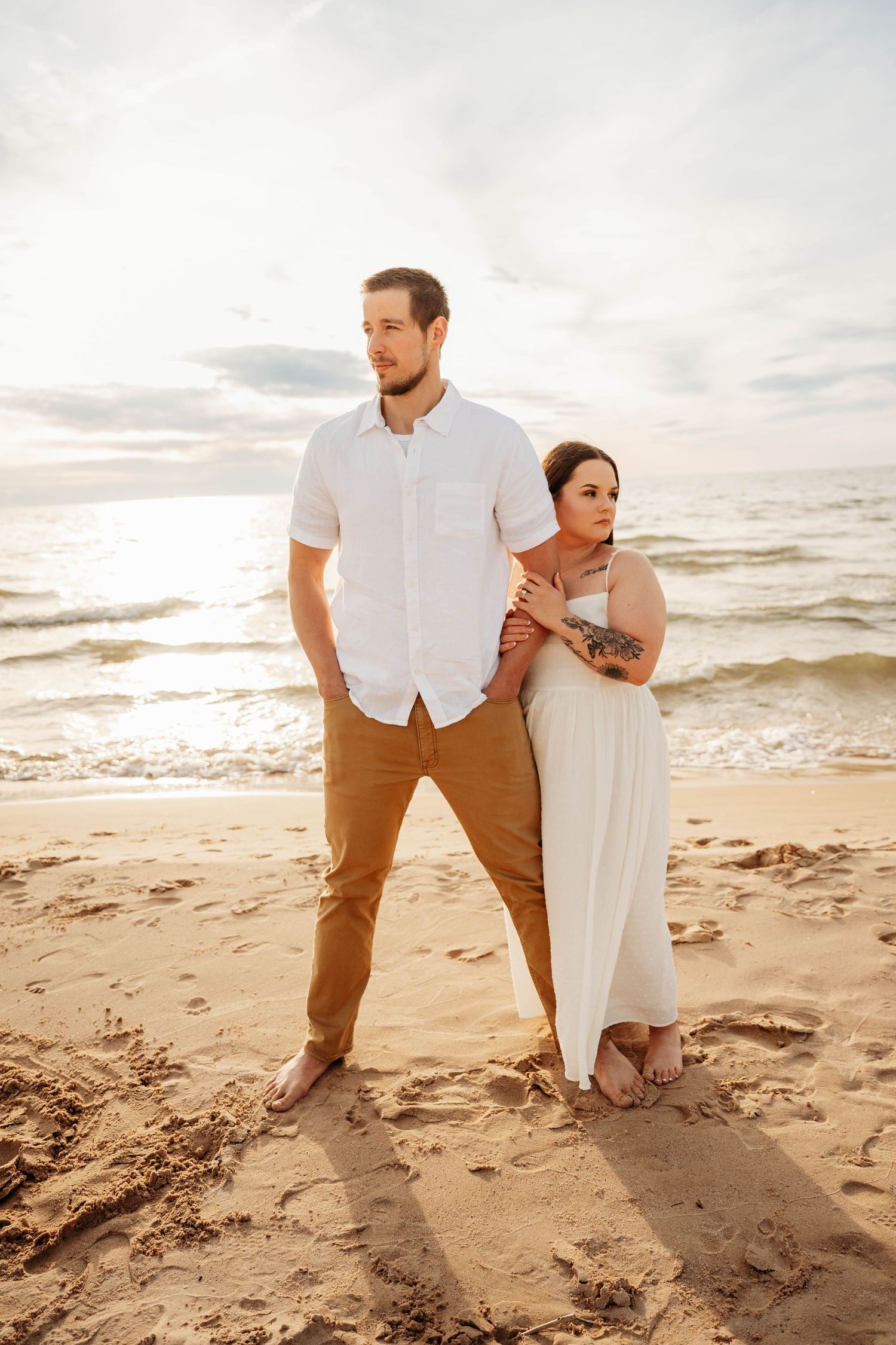 Couple standing together on beach at sunset, woman in white dress embracing man.