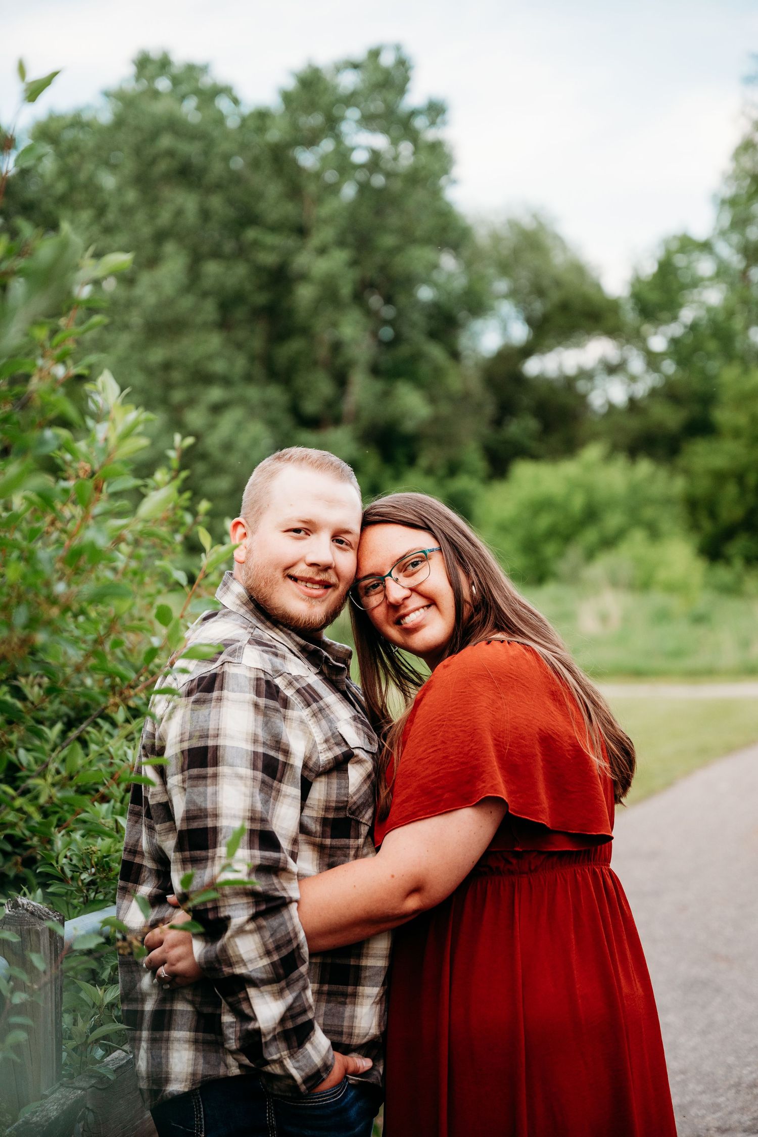 A smiling couple embraces outdoors, woman in red dress leans on man in plaid shirt, surrounded by lush green trees.