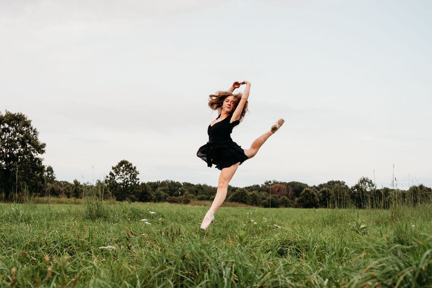 Ballet dancer gracefully leaps across a green meadow during a summer afternoon performance.