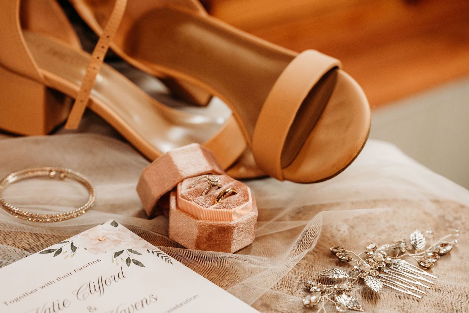 Wedding flat lay featuring tan heels, velvet ring box with gold bands, bridal hair comb, bracelet, and floral invitation on tulle.