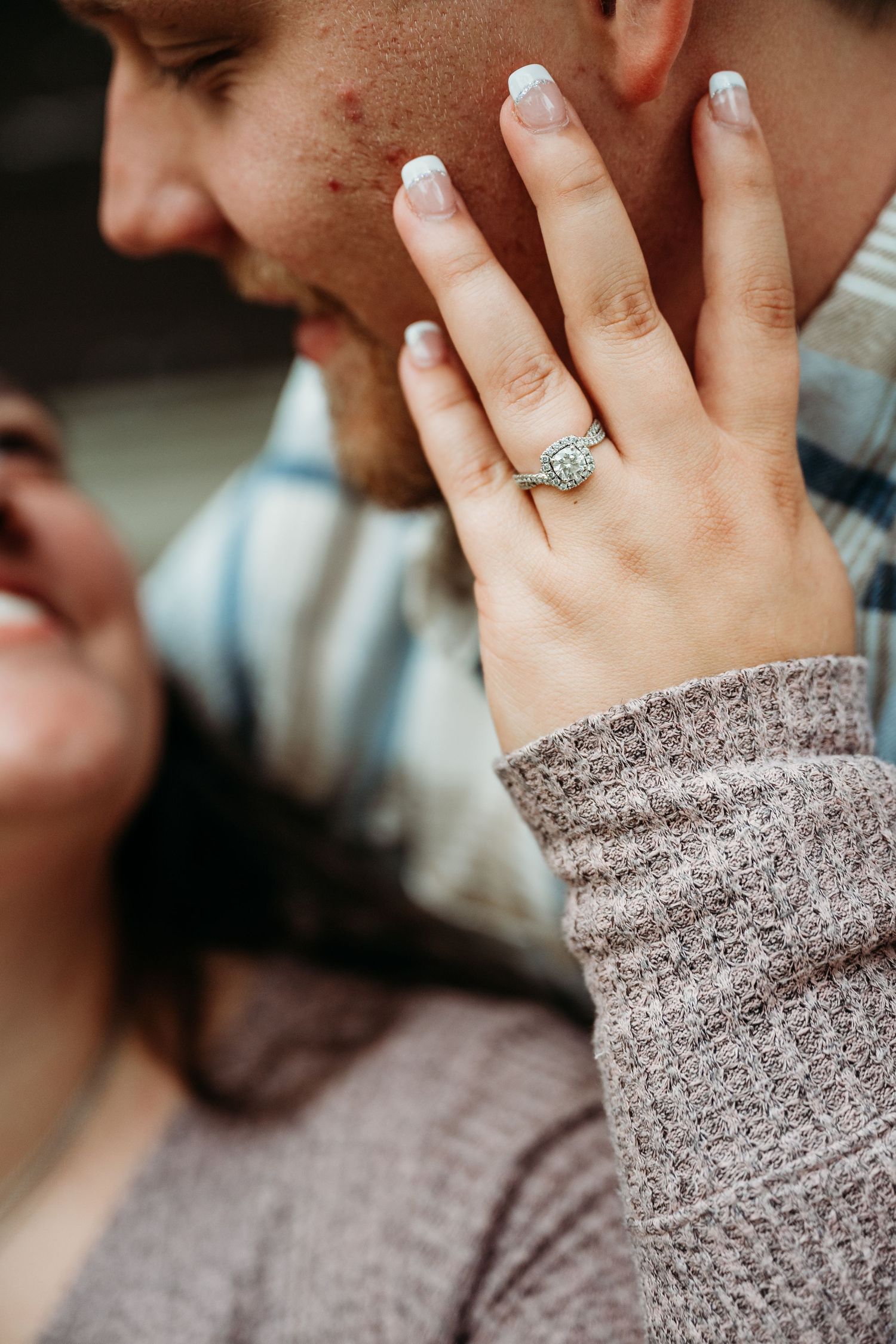 Close-up view of a hand wearing a sparkling diamond engagement ring against a gray knit sweater sleeve.