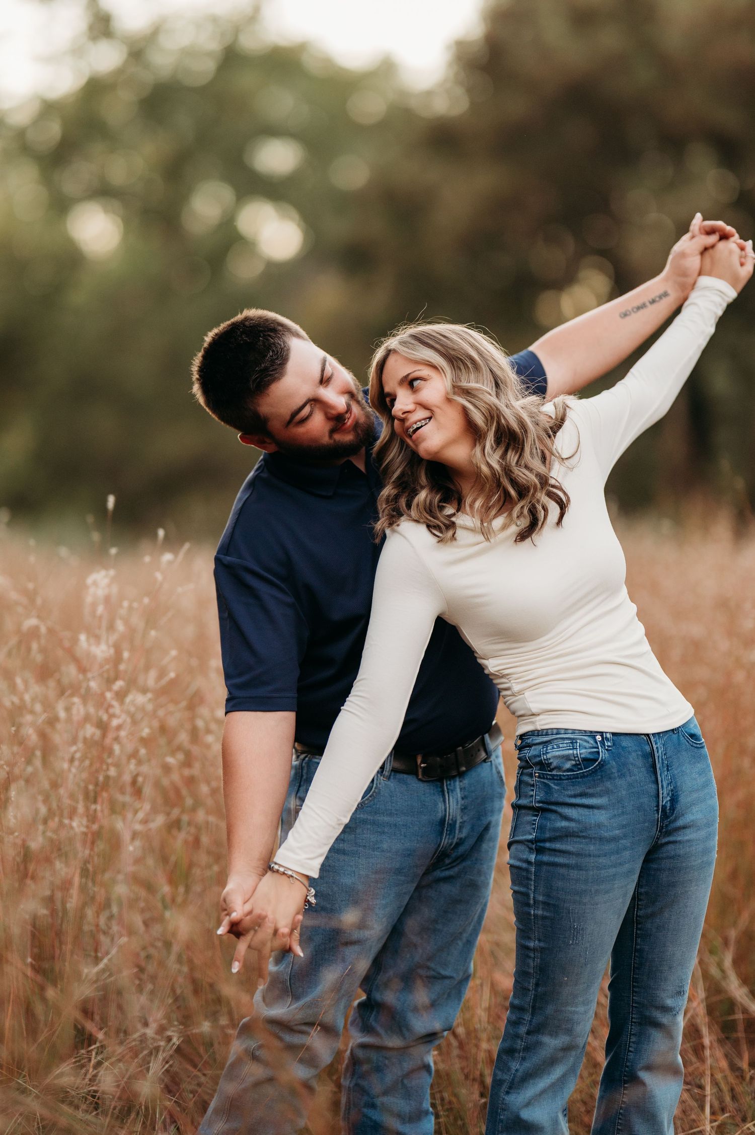 A couple shares playful moments in a golden field at sunset, dressed casually in jeans and white tops.