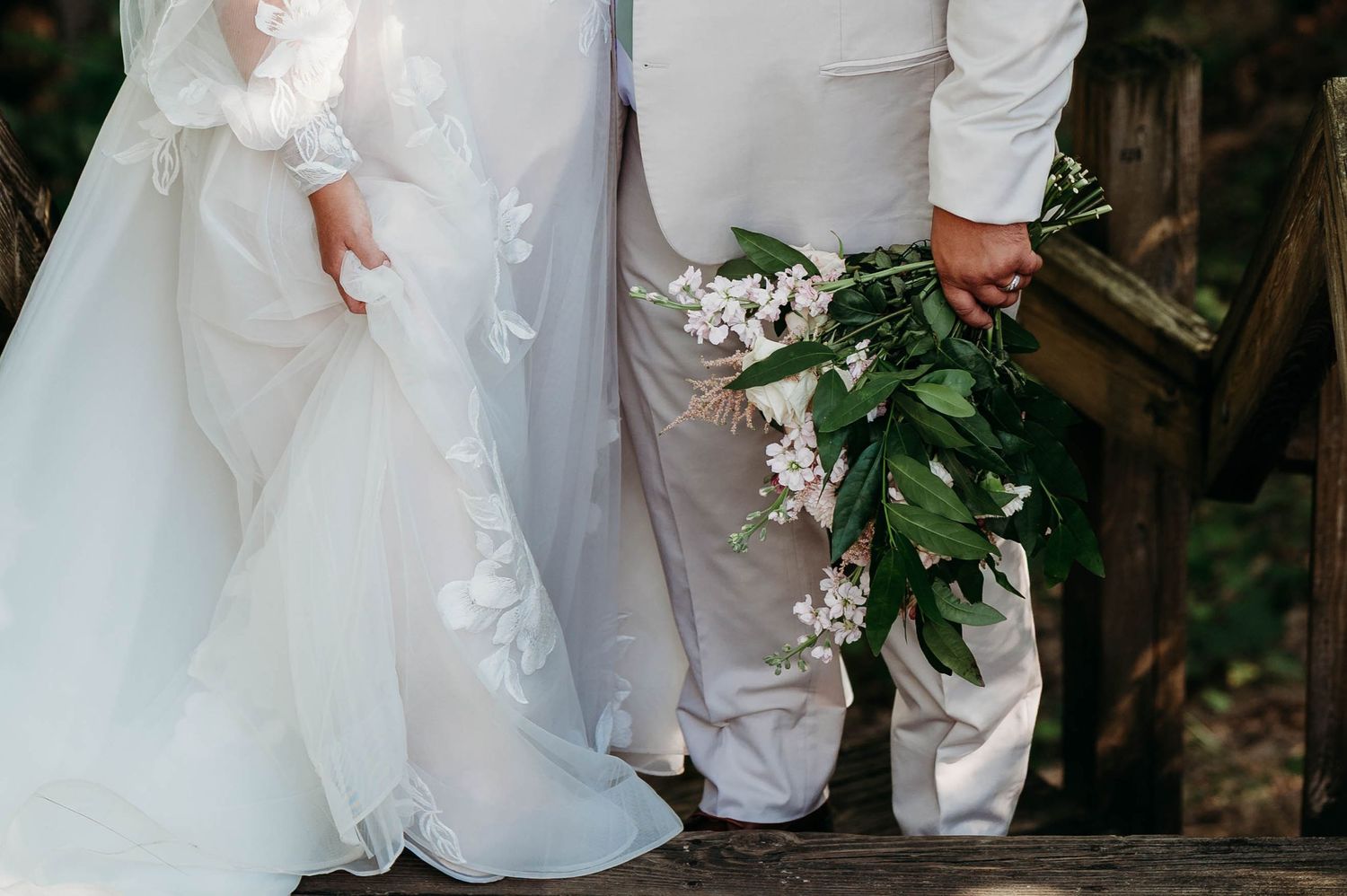 A couple in wedding attire stands on wooden steps, with white flowers and greenery in their hands.