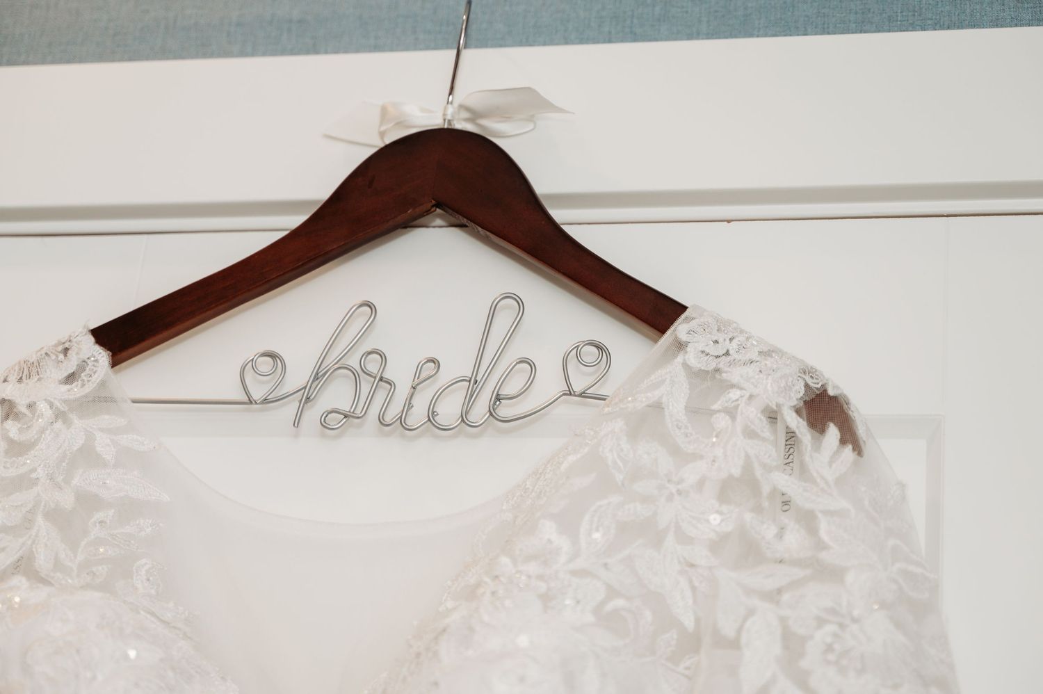 White wedding dress hangs on a wooden hanger with the word 'bride' written in silver wire above it.