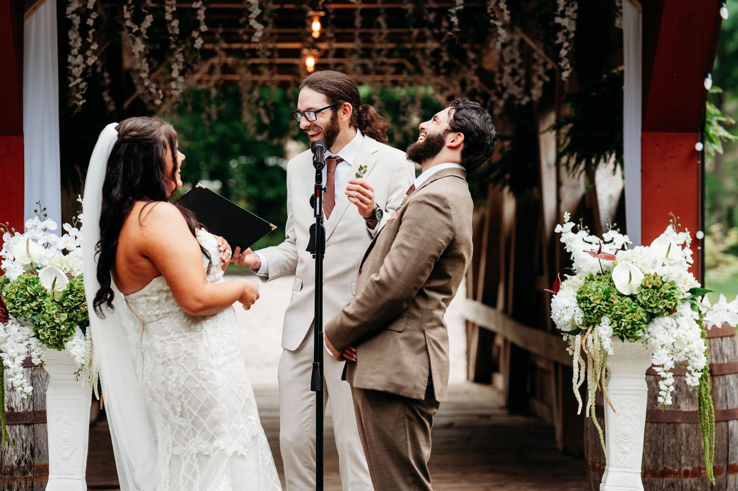 Wedding ceremony with bride, groom, and officiant laughing under a rustic pavilion with white floral arrangements.