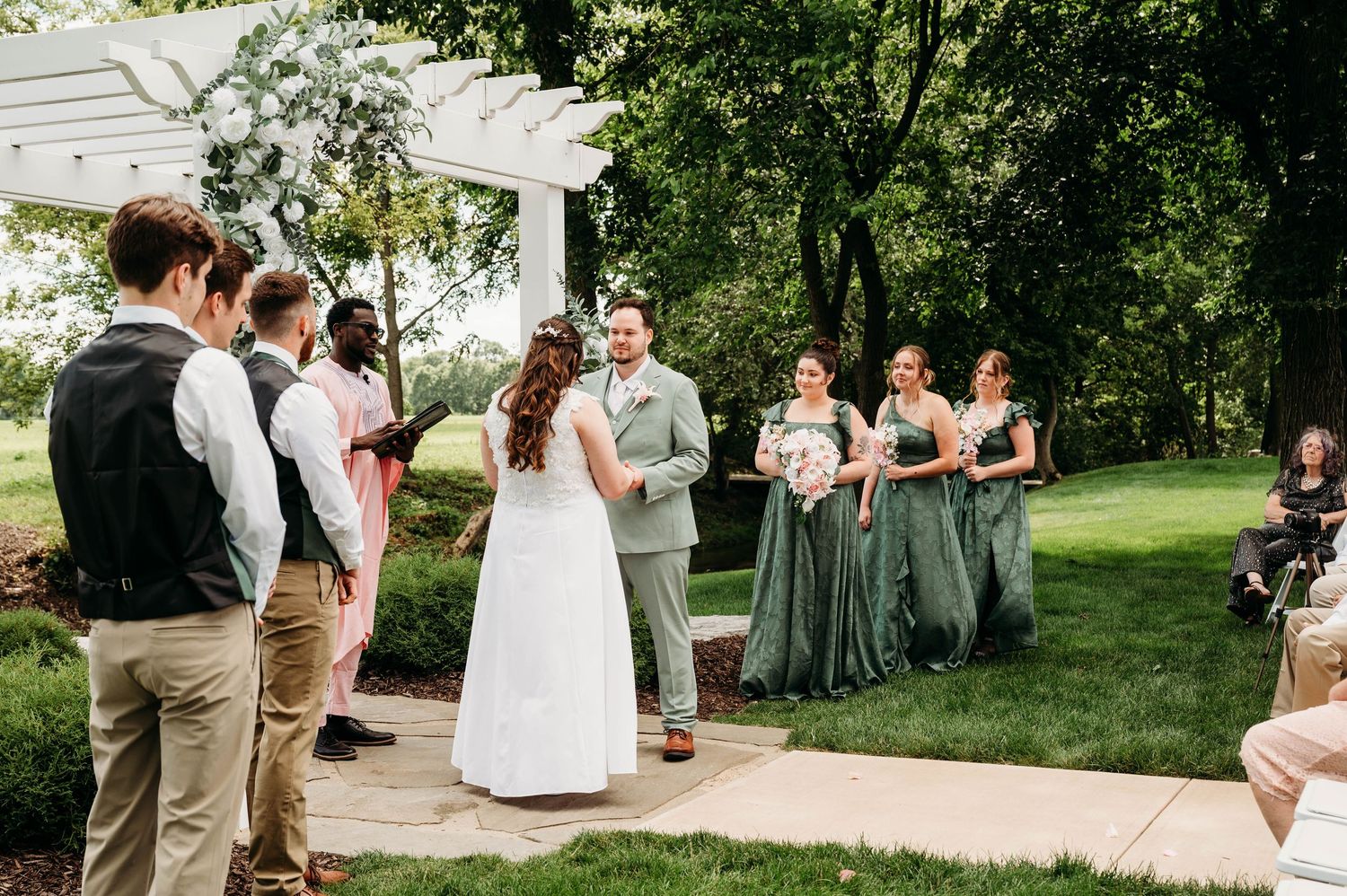 Outdoor wedding ceremony on a patio with bride and groom facing each other, bridesmaids in sage green dresses, lush green trees.