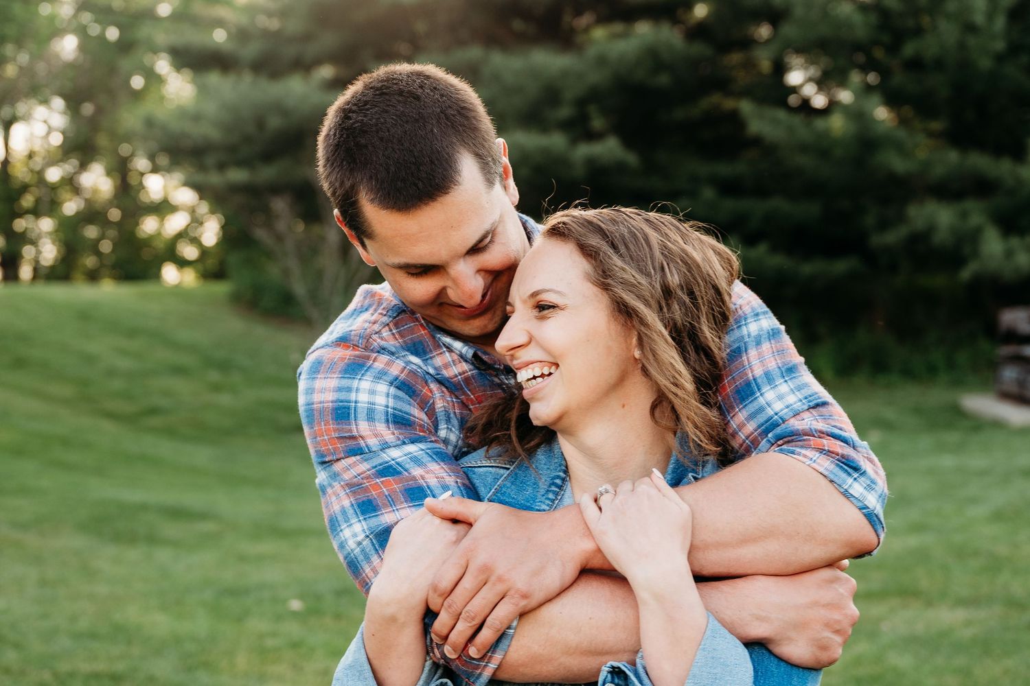 Happy couple embracing outdoors, man hugging woman from behind as she smiles joyfully in a sunny green park.